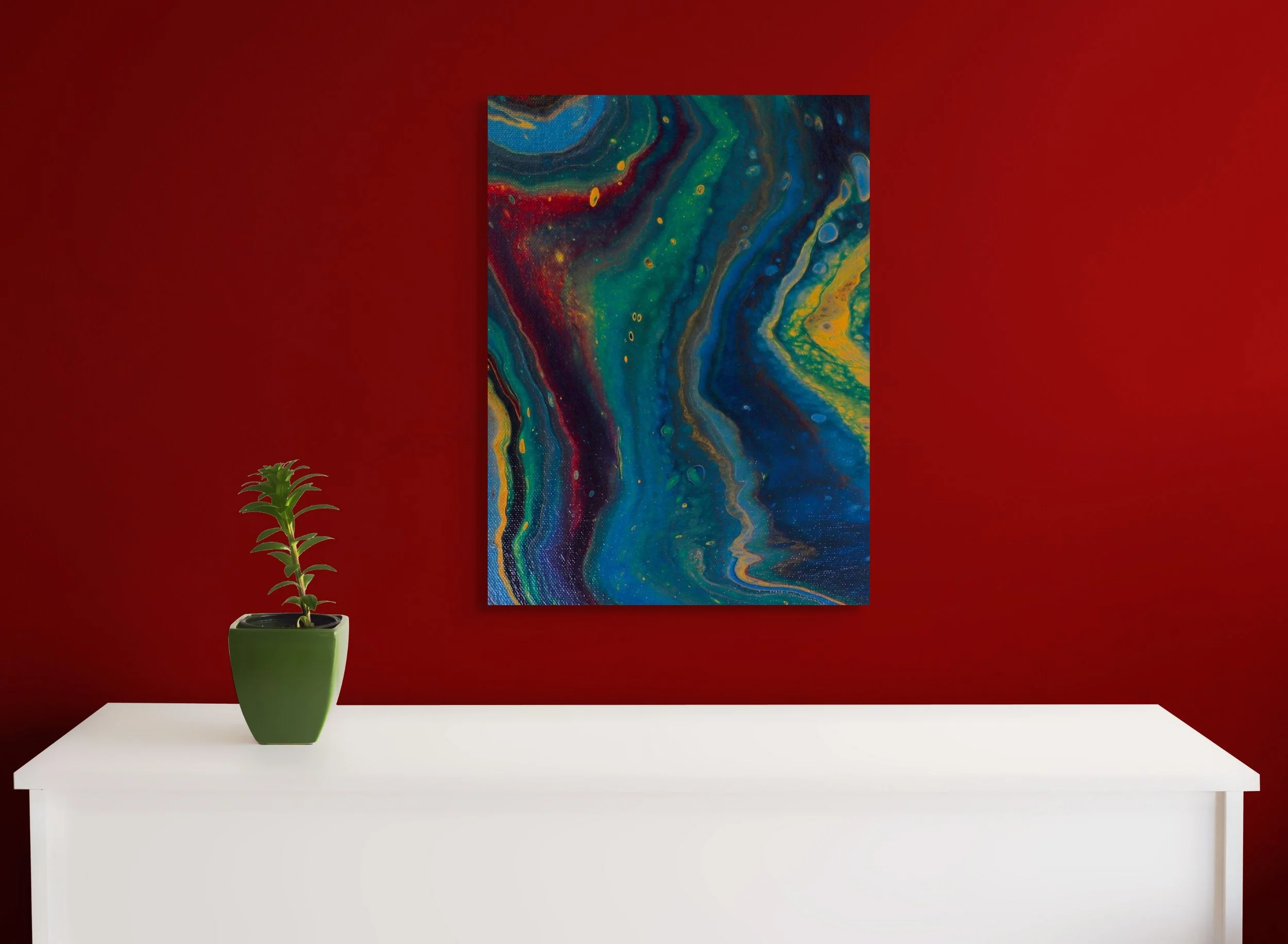 An interior setting with a red wall, a colorful abstract painting hanging on it, a white table, and a green potted plant on top of the table.