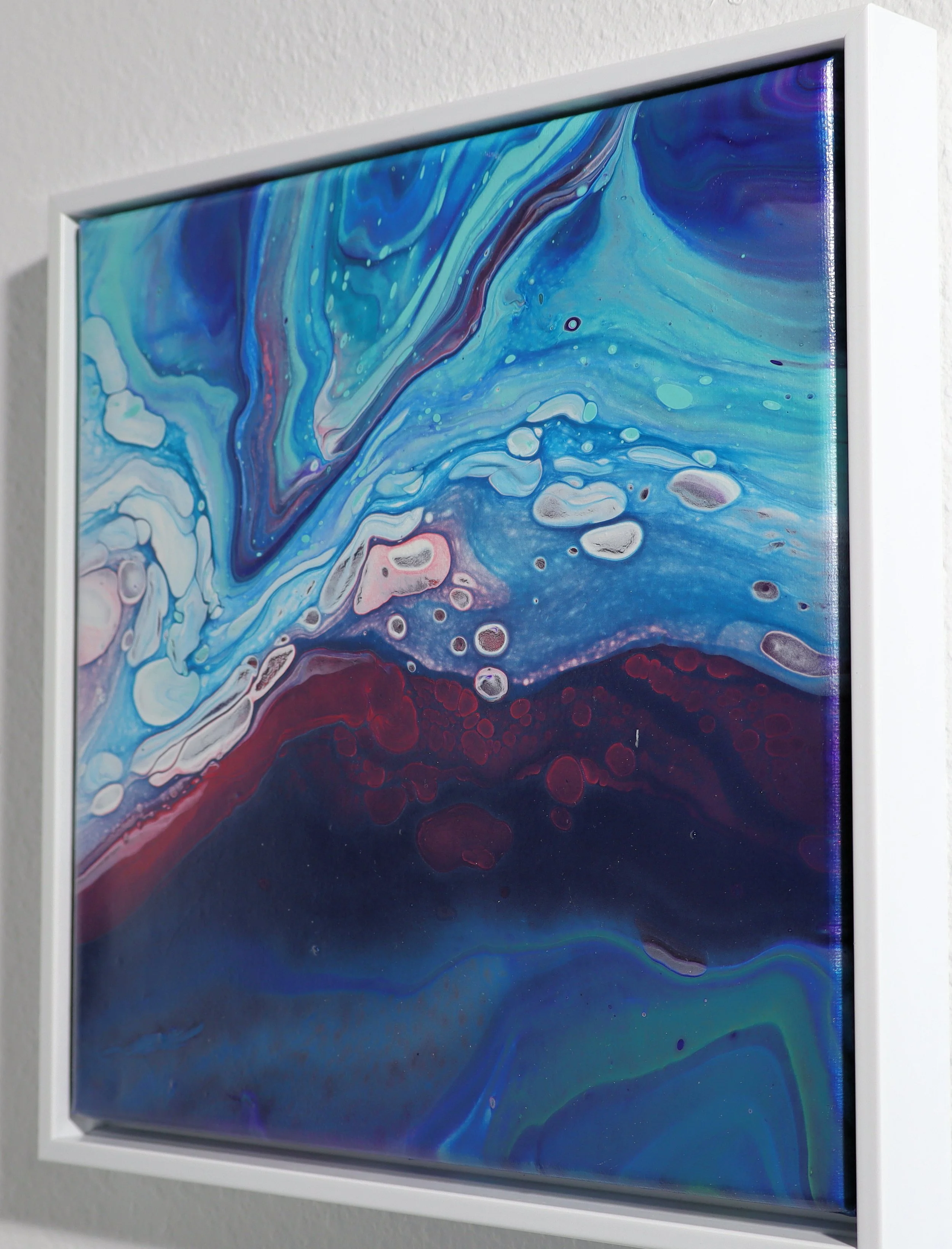 Framed abstract painting with swirling patterns of blue, purple, and pink on a white wall.