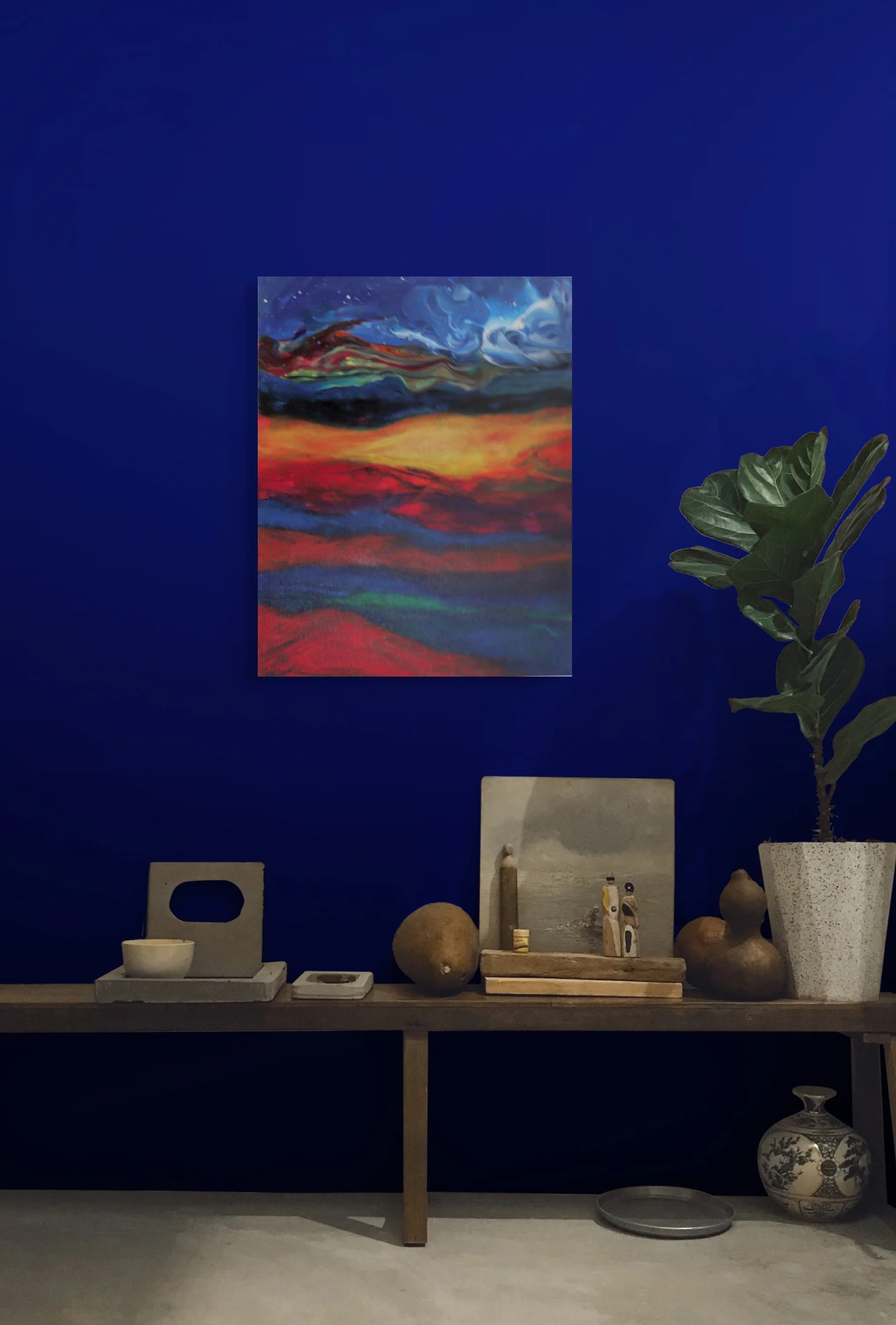A display wall with a large colorful abstract painting, a wooden table with various pottery and art objects, a tall potted fig plant in a textured white pot, and additional artwork and decorative items on and below the table, set against a dark blue 