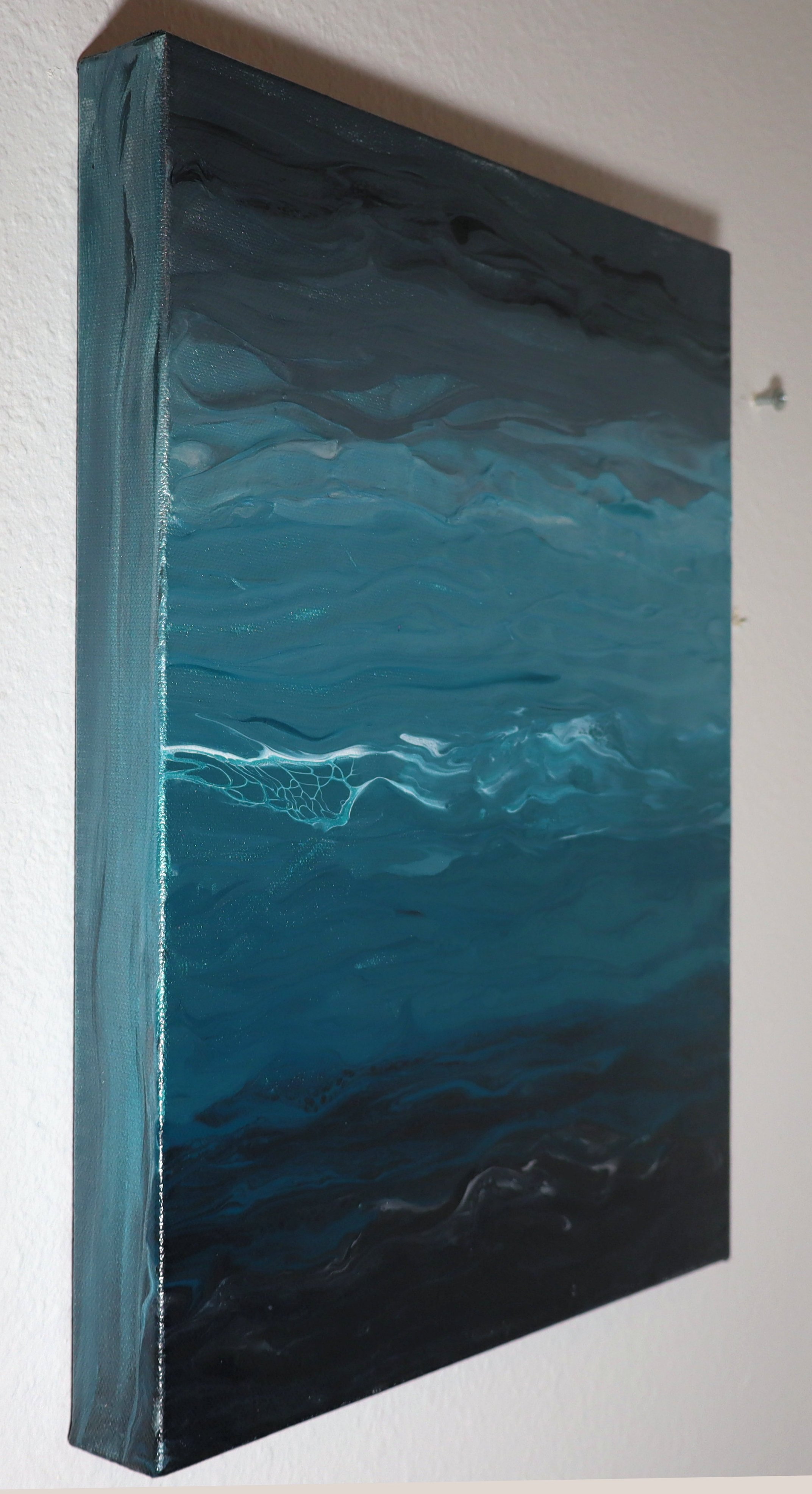 A vertical abstract acrylic painting with swirling shades of blue, black, and gray on a canvas, hanging on a white wall.
