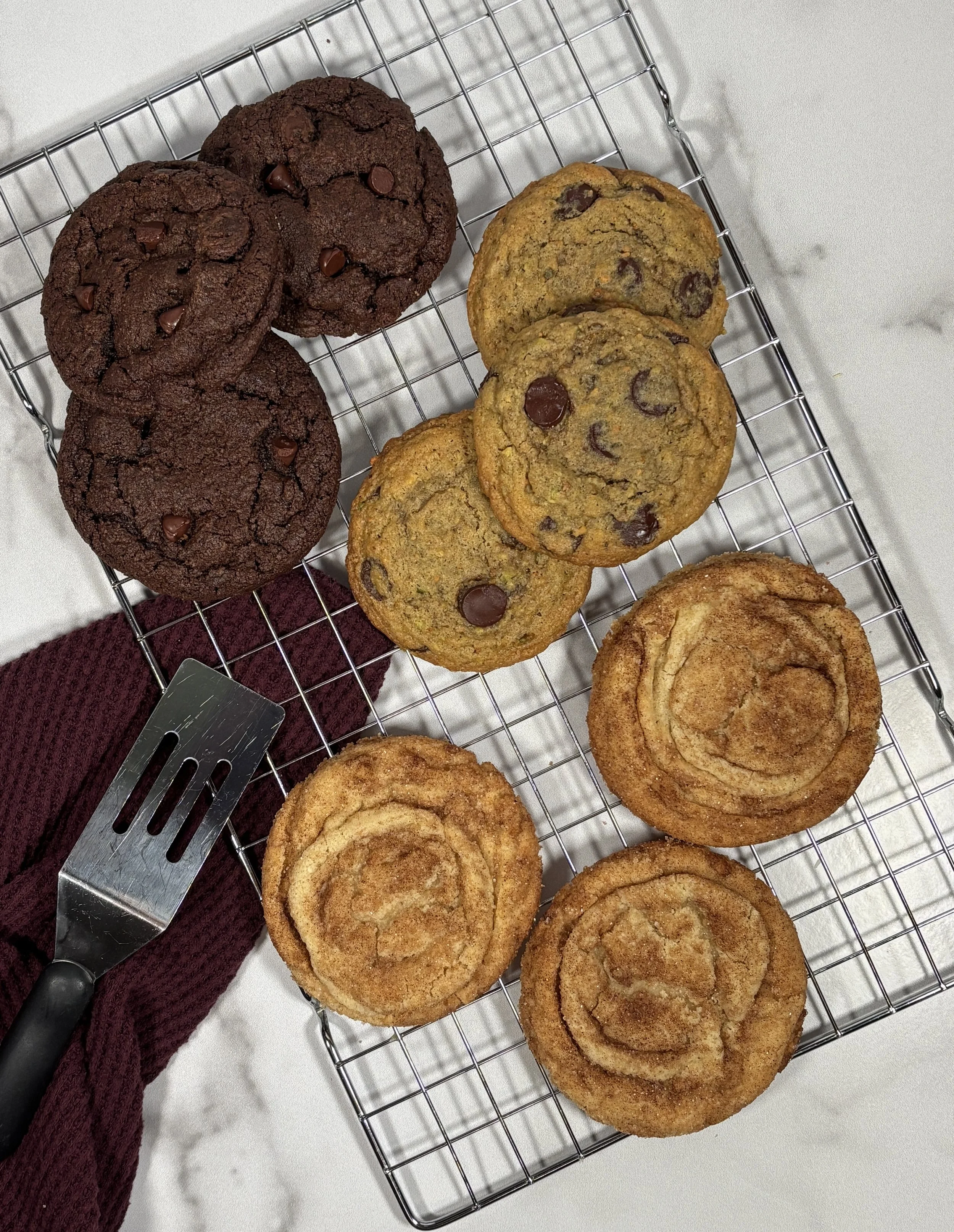 Chocolate chip cookies and cinnamon rolls cooling on a wire rack on a white marble surface, with a burgundy cloth and a metal spatula nearby.