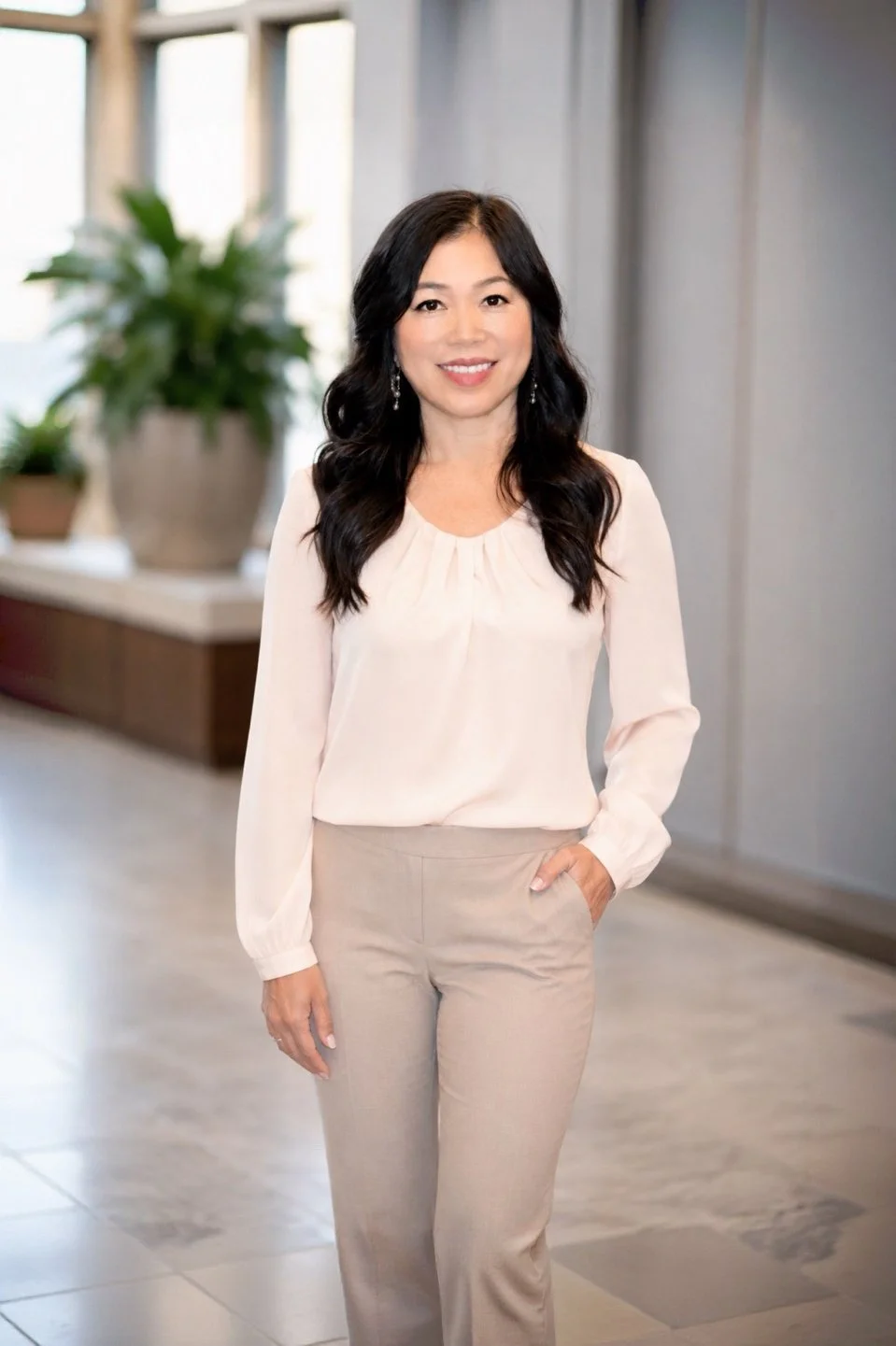 A woman with long black hair wearing a light pink blouse and beige pants standing indoors near a window with potted plants in the background.