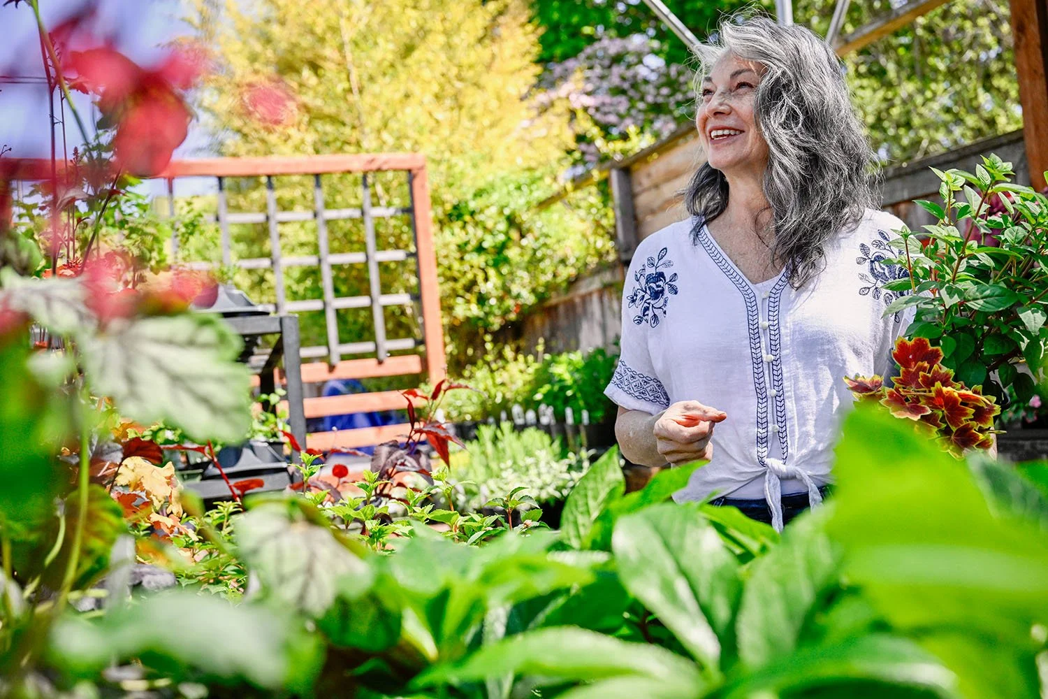 Kellie Torres of Portland, smiling and standing in a garden filled with various green and colored plants, with a wooden fence and garden furniture in the background on a sunny day.