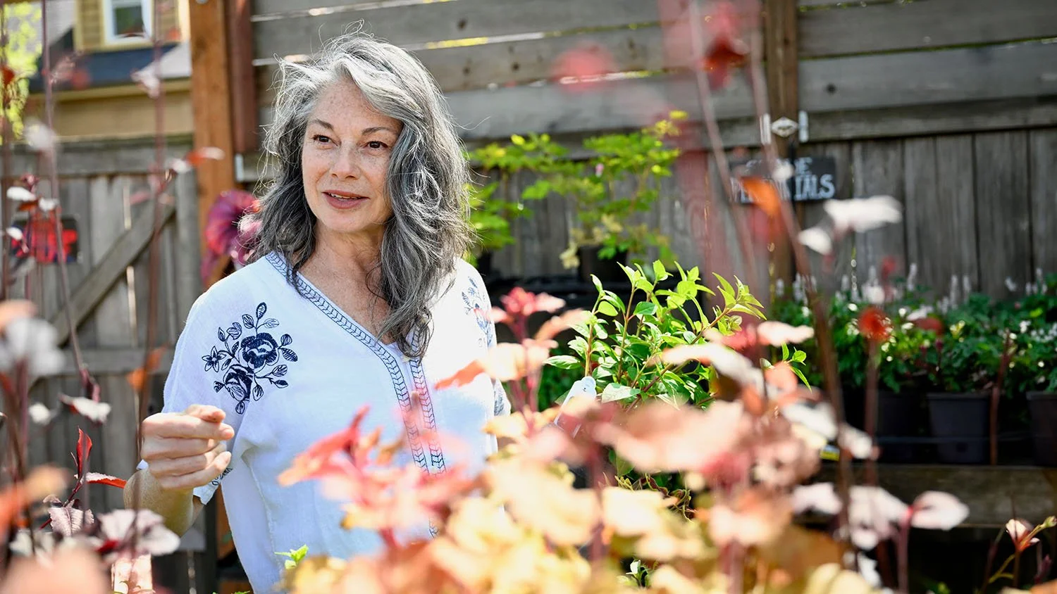 Kellie Torres stands outdoors in a garden filled with pink, red, and green plants, with a wooden fence and a small window in the background.
