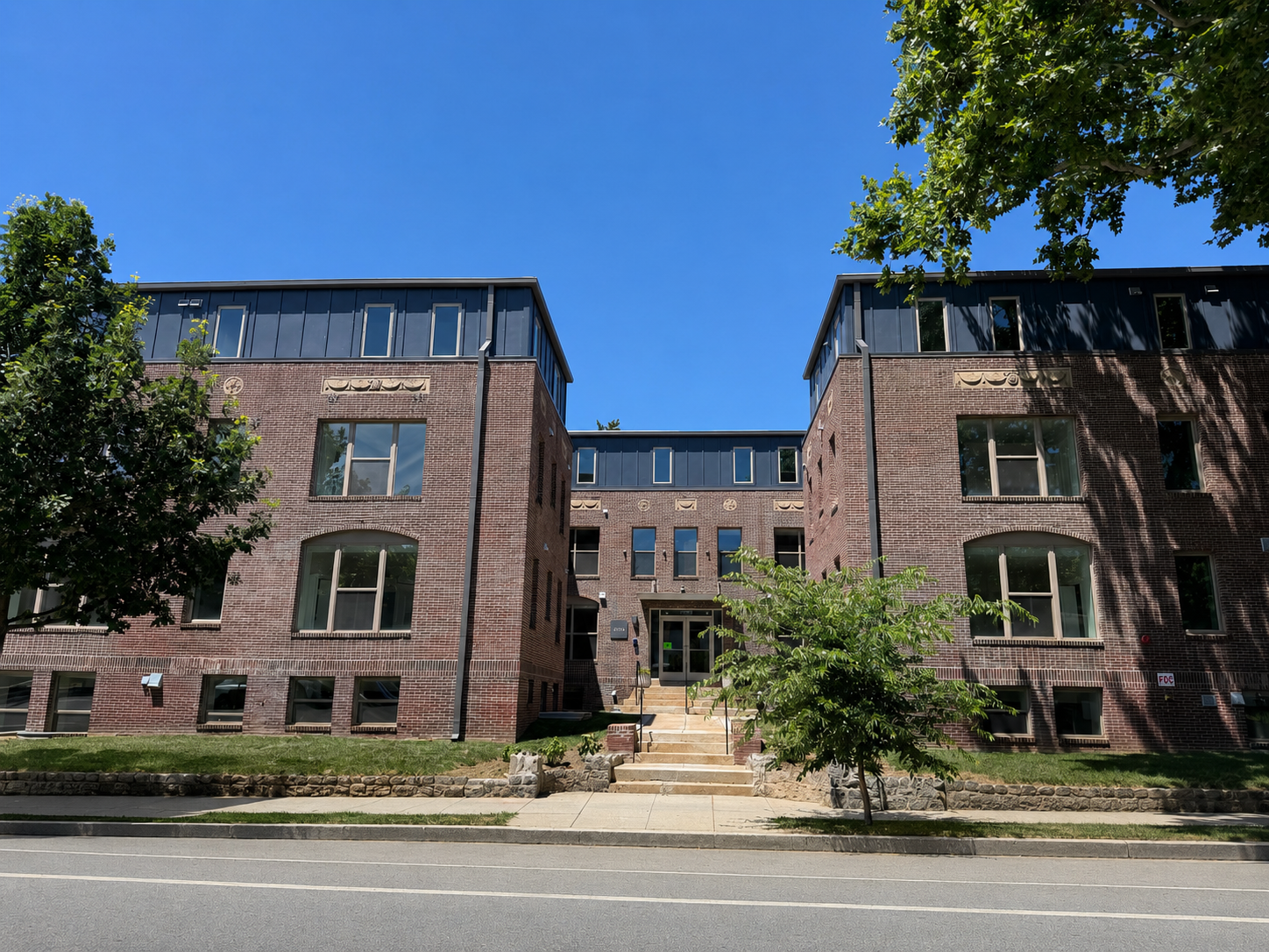A three-story brick building with black trim and a central entrance accessed by stairs, surrounded by trees and greenery, under a clear blue sky.