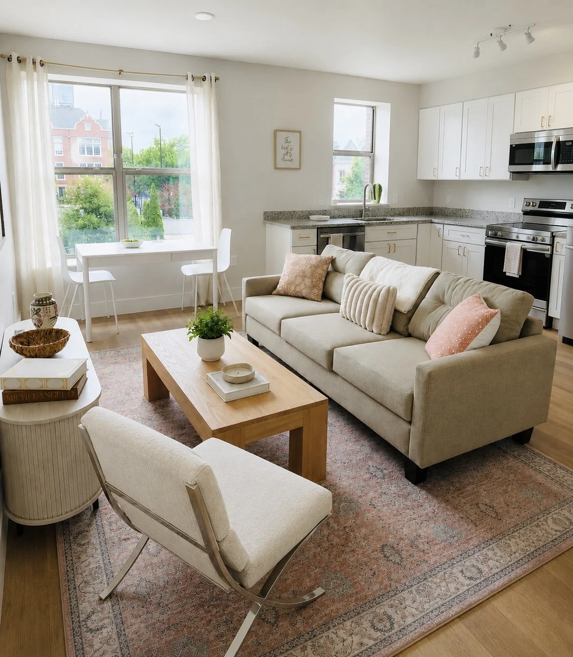 Bright, open-concept living room and kitchen with large windows, white cabinets, gray countertops, a beige sofa with pillows, a wooden coffee table, a patterned area rug, and a small dining area with a white table and chairs.