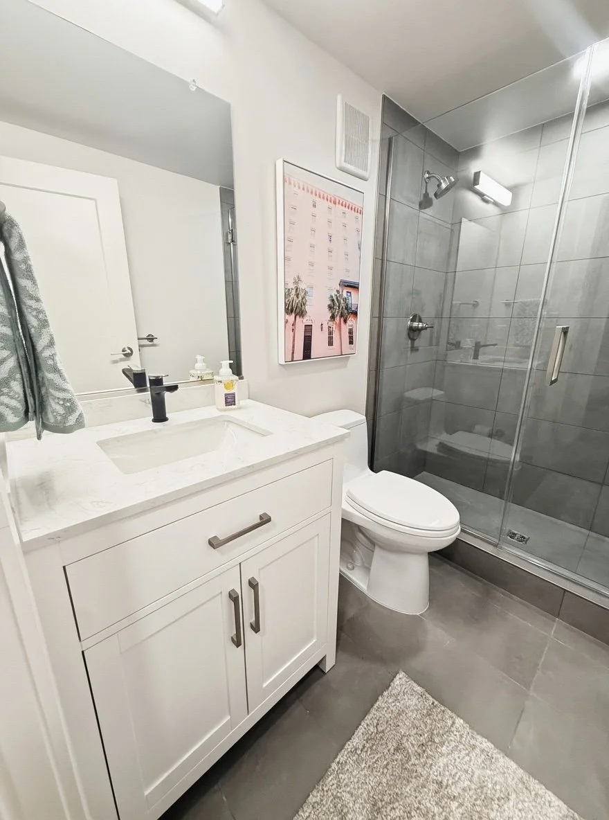 Modern bathroom with white vanity, marble countertop, black faucet, soap dispensers, toilet, glass-enclosed shower with gray tiles, wall-mounted showerhead, and a beige rug.