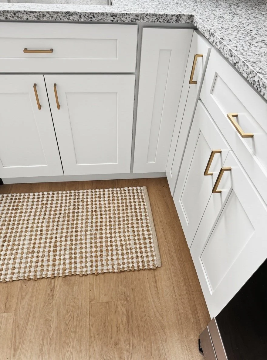Close-up of white kitchen cabinets with gold handles, granite countertop, and a beige patterned rug on wooden flooring.