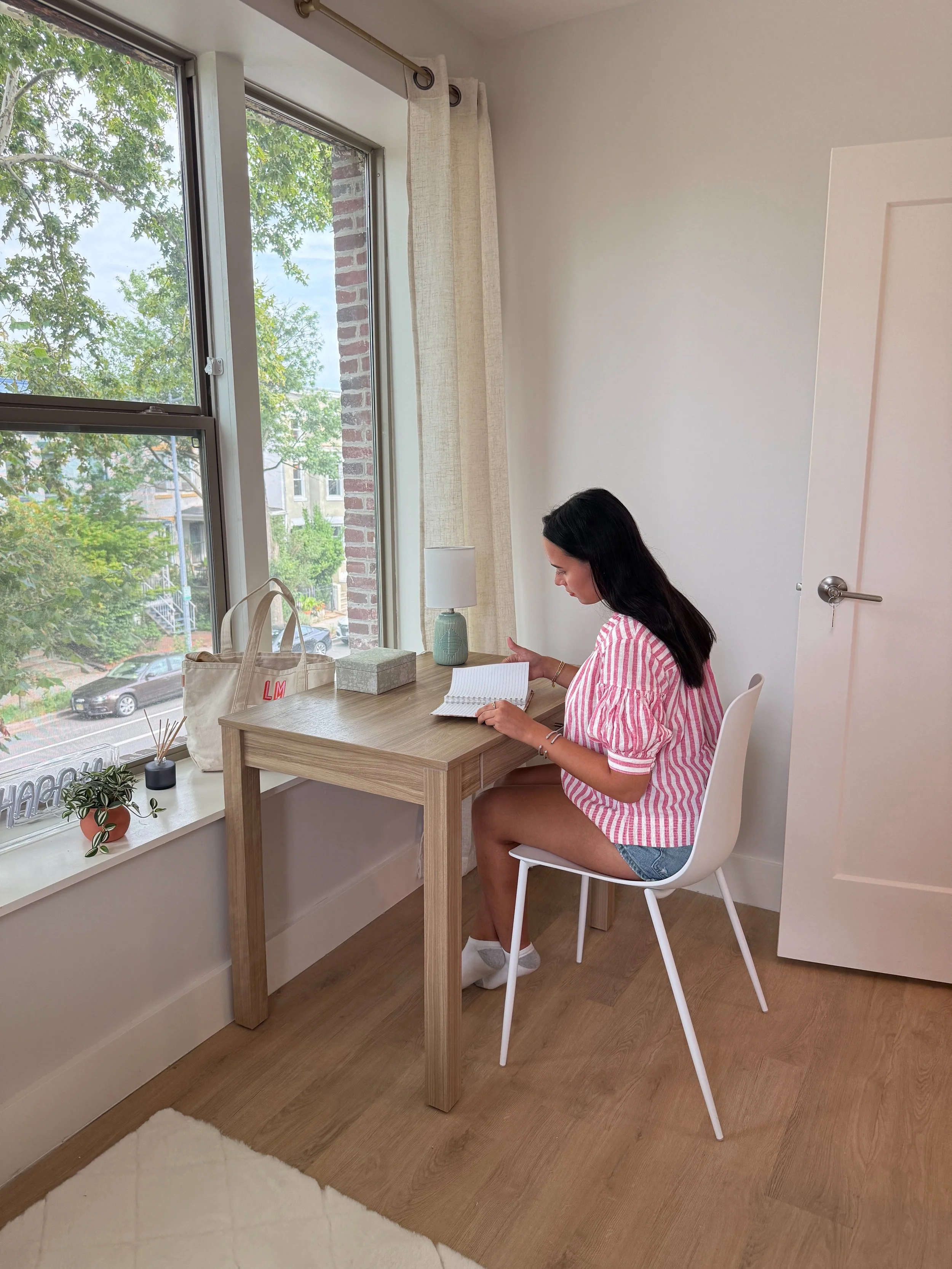 A woman with long dark hair sitting at a small wooden desk in front of a window with beige curtains. She is reading a notebook. The room has white walls, a lamp, and a beige tote bag on the desk. Outside the window, trees and buildings are visible.