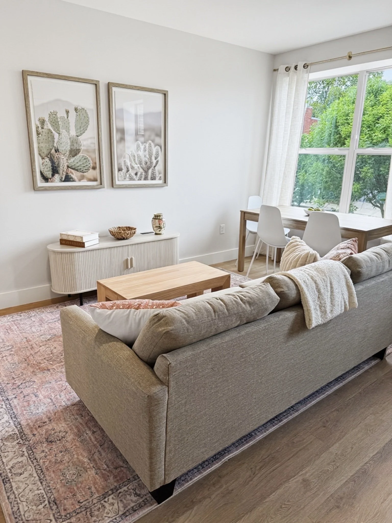 A cozy living room with a beige sofa, a wooden coffee table, a white sideboard with decorative items, and wall art featuring framed cactus prints. There's a large window with white curtains letting in natural light and outdoor greenery visible outsid