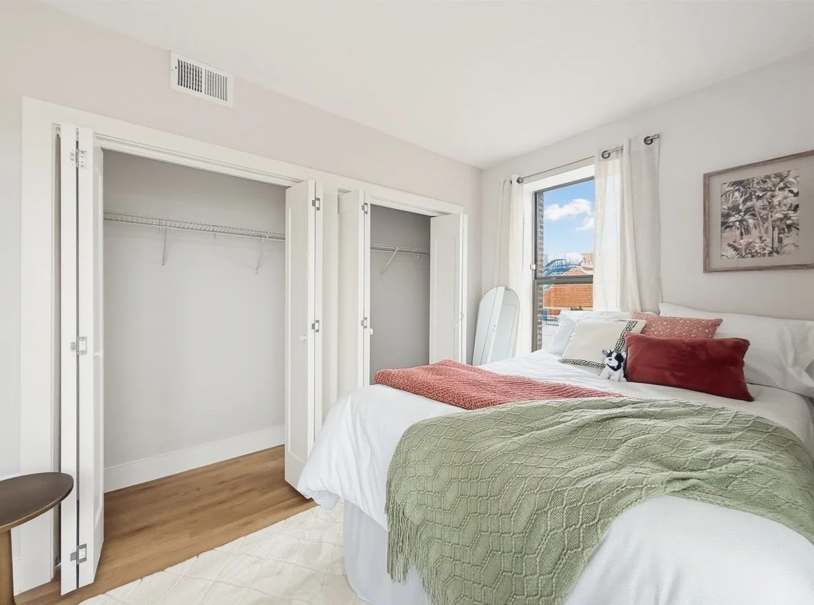 A bedroom with a bed, pillows, a blanket, a mirror, a window with curtains, and open closet doors.