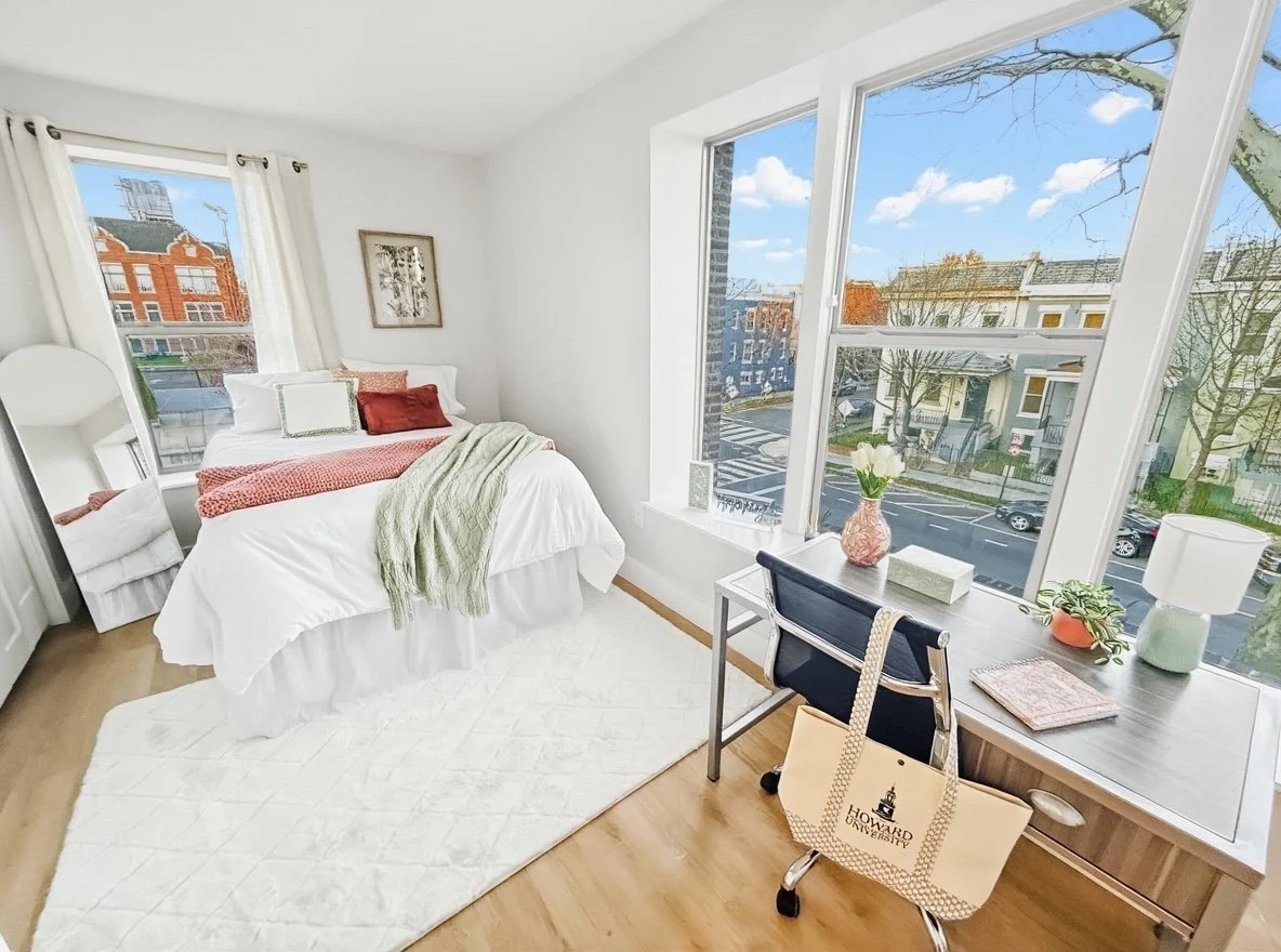Bright bedroom with a white bed, green throw blanket, and decorative pillows, next to large windows showing a street view with houses and trees, a white rug on wooden floor, and a desk with a chair, vase with flowers, and a bag with Howard University