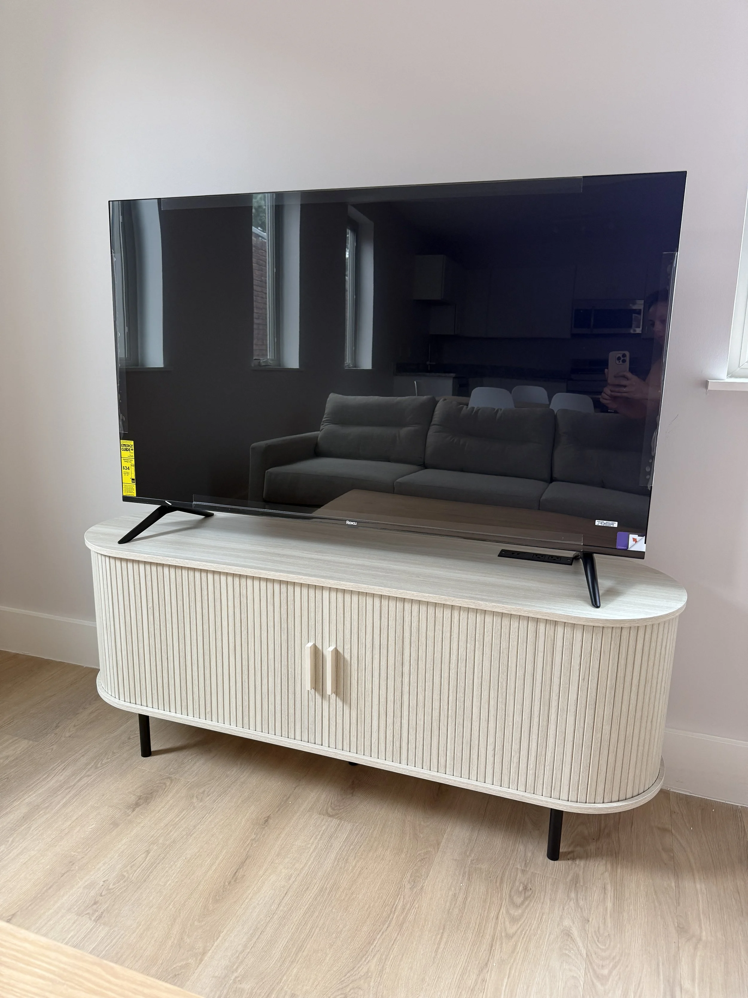 A flat-screen television on a white wooden stand with black legs, placed on a light-colored wooden floor, with a reflected living room visible on the screen including a gray sofa, dining table, and kitchen with cabinets.