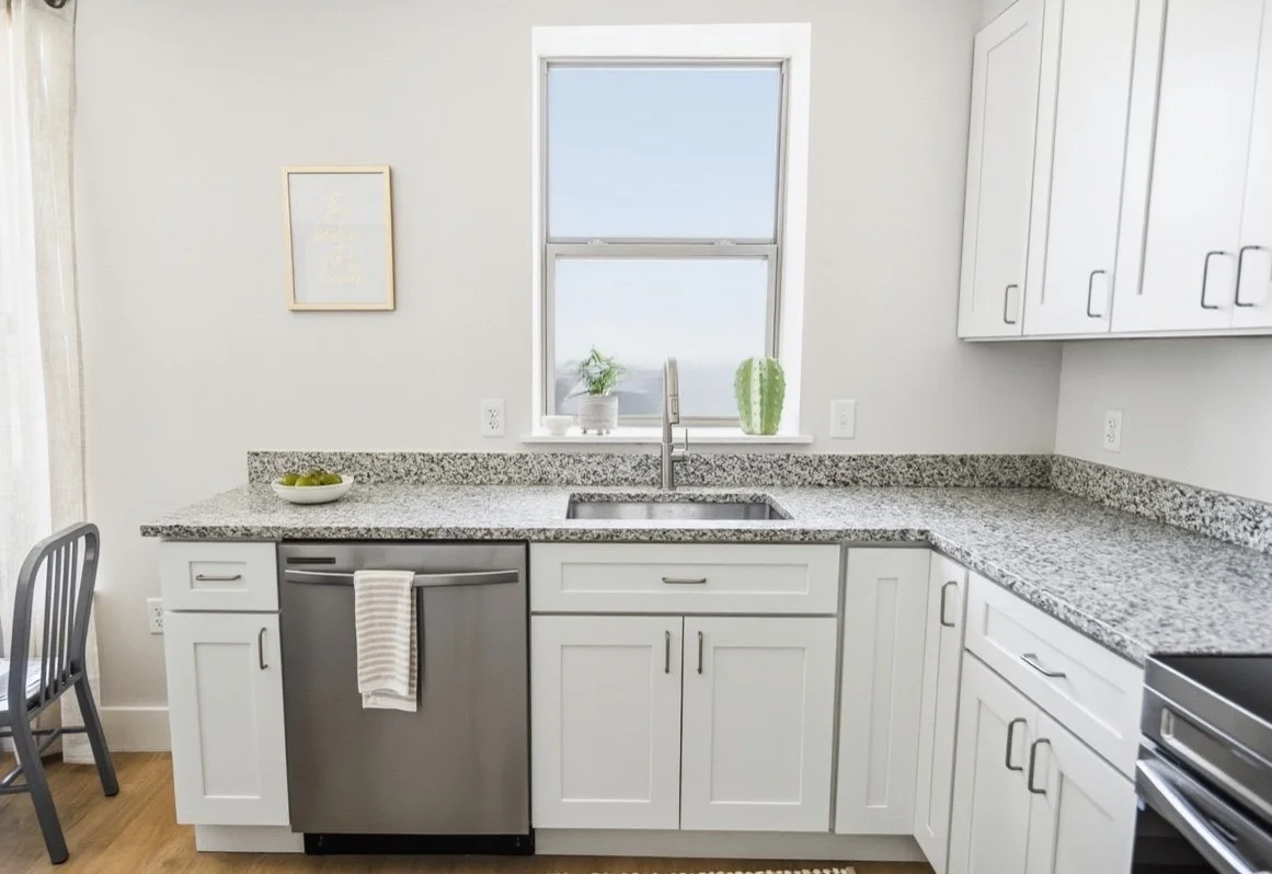 Kitchen with white cabinets, granite countertops, and a window above the sink. There are potted plants on the windowsill, a bowl of fruit on the counter, and a dishwasher with a towel hanging on it.