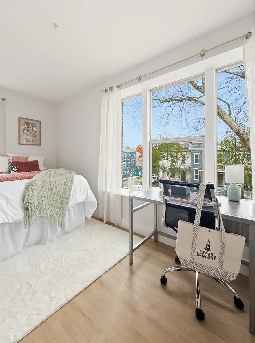 A bright bedroom with a bed, desk, and window showing trees and a building outside.