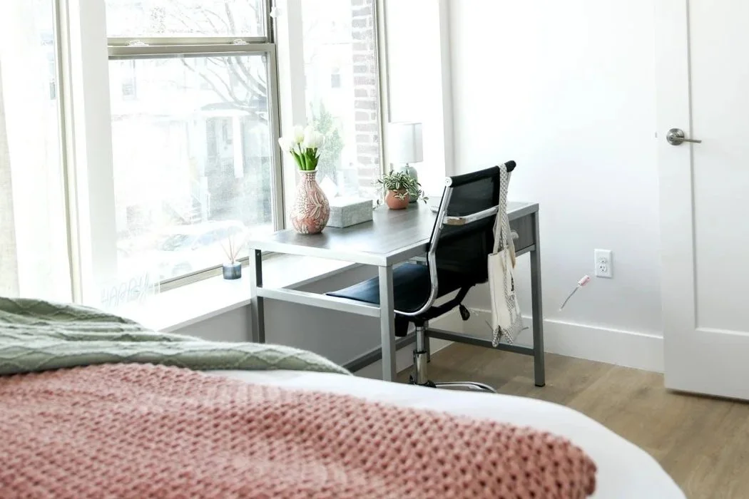 A bedroom corner with a bed in the foreground, a desk near a large window with flower vases, a potted plant, and a lamp.