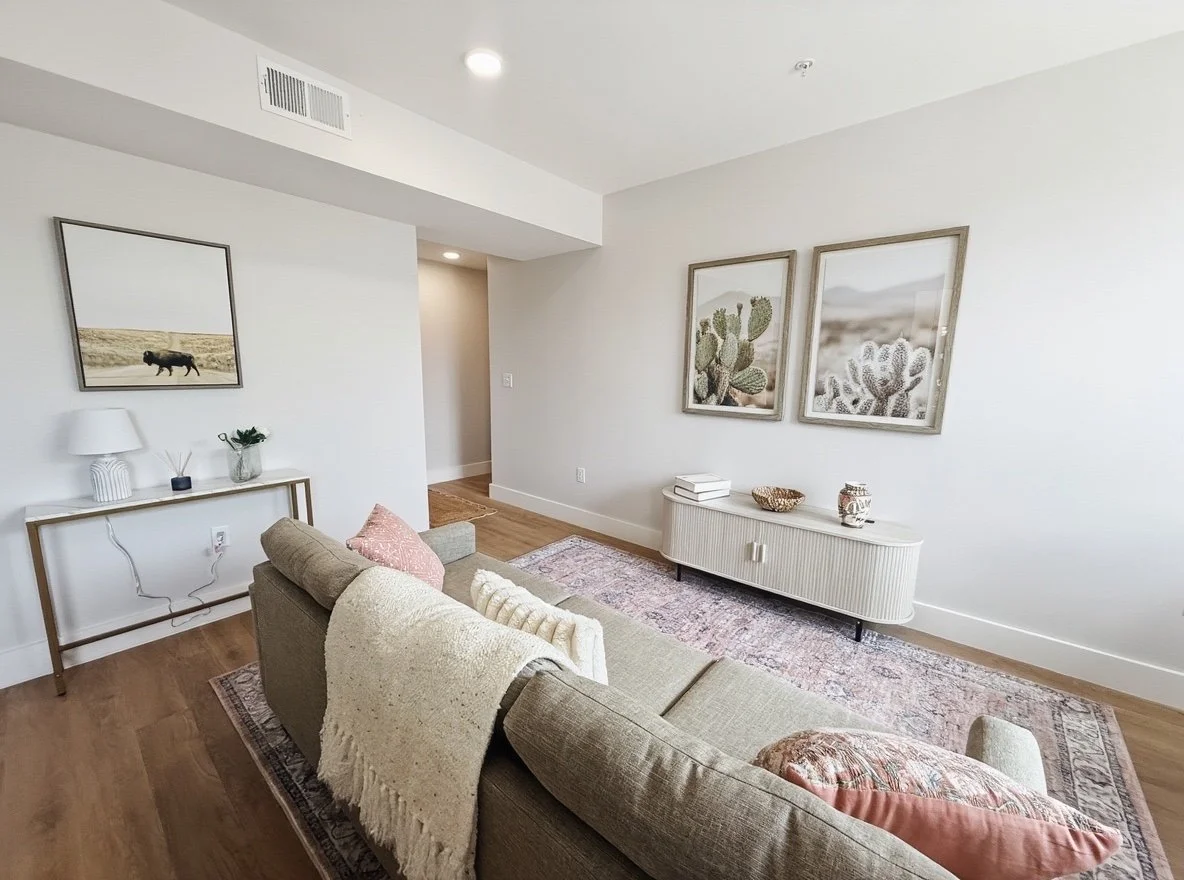 Living room with beige couch, pink and cream pillows, white sideboard, wall art of desert scenes and cacti, hardwood floor, and decorative items.