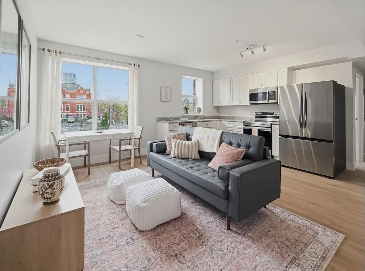 A bright living room with a black sofa, patterned pillows, a white blanket, and two white ottomans on a pink rug. Behind the sofa is a kitchen with white cabinets, a stainless steel refrigerator, stove, and microwave. To the left, a large window with