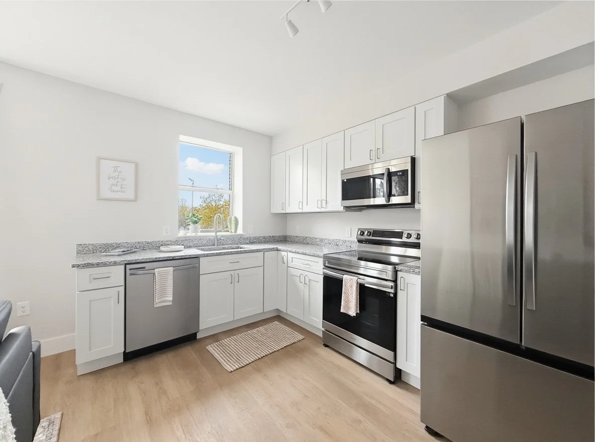 Modern kitchen with white cabinets, stainless steel appliances including a refrigerator and oven, granite countertops, a window above the sink, and a light wood floor.