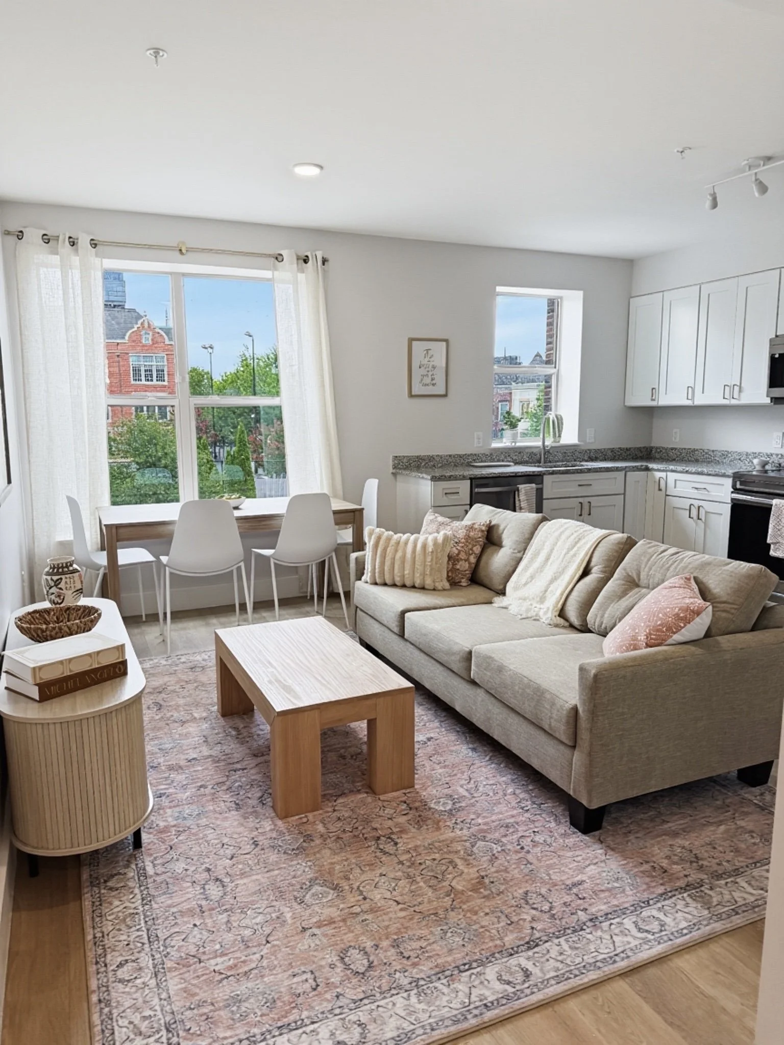 A living room with a beige sofa, patterned rug, coffee table, and dining table with six chairs near large windows with white curtains. The kitchen has white cabinets, granite countertops, and a window above the sink with a city view.