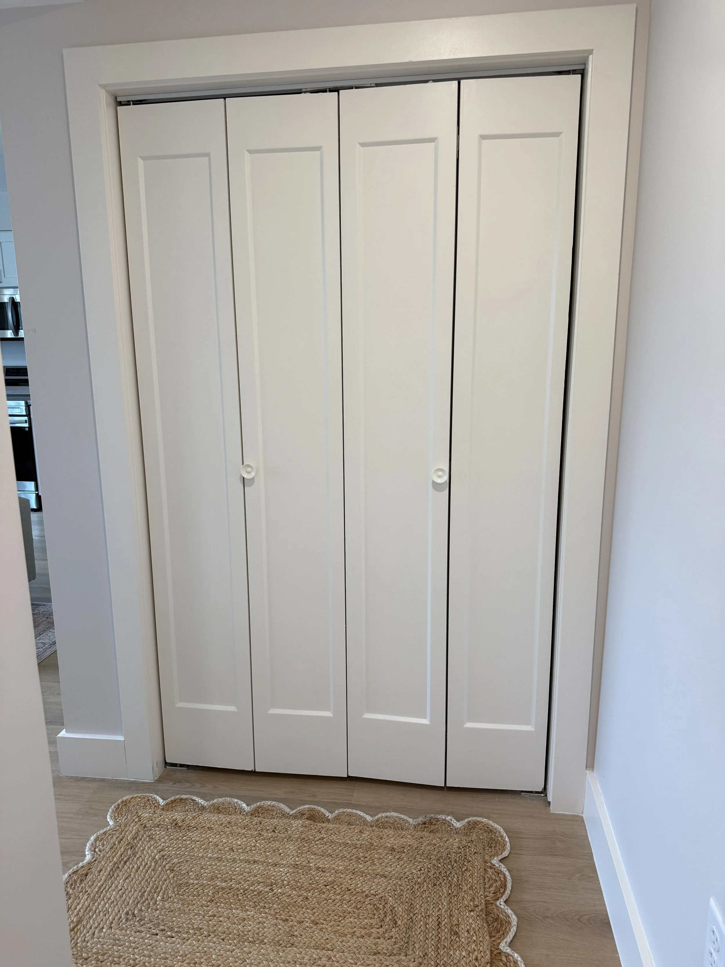 White closet with four folding doors and round white knobs. Brown woven rug with scalloped edges on light wood floor in entryway. Part of kitchen visible on the left.