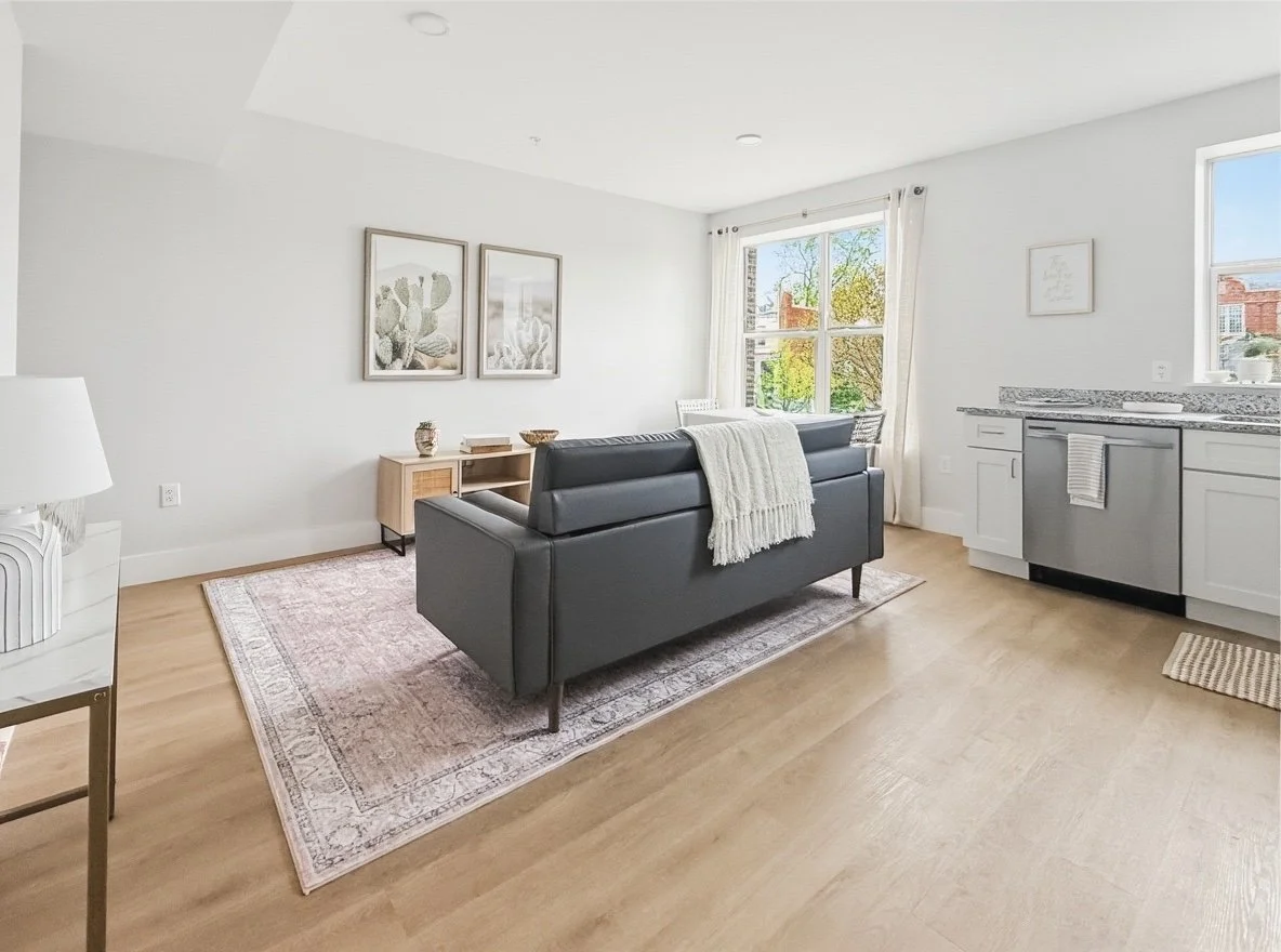 Living room with a black sofa, a small wooden side table, framed cactus artwork on the wall, a white and beige patterned rug, and large windows with white curtains letting in natural light. The adjacent area has a compact kitchen with white cabinets 
