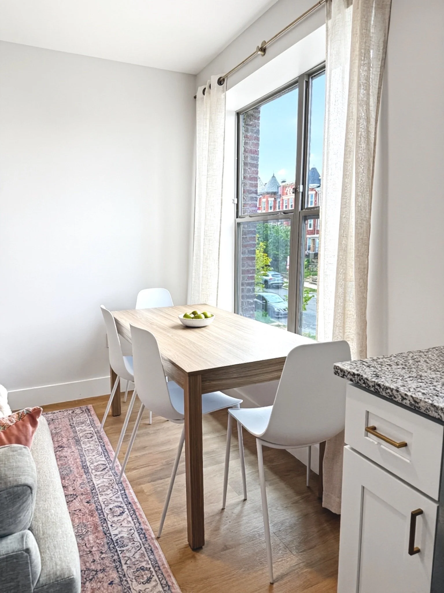 A dining area with a wooden table, four white chairs, a bowl of green apples, large window with curtains, view of buildings outside, hardwood floor, part of a gray sofa, and a white cabinet with gold handles.
