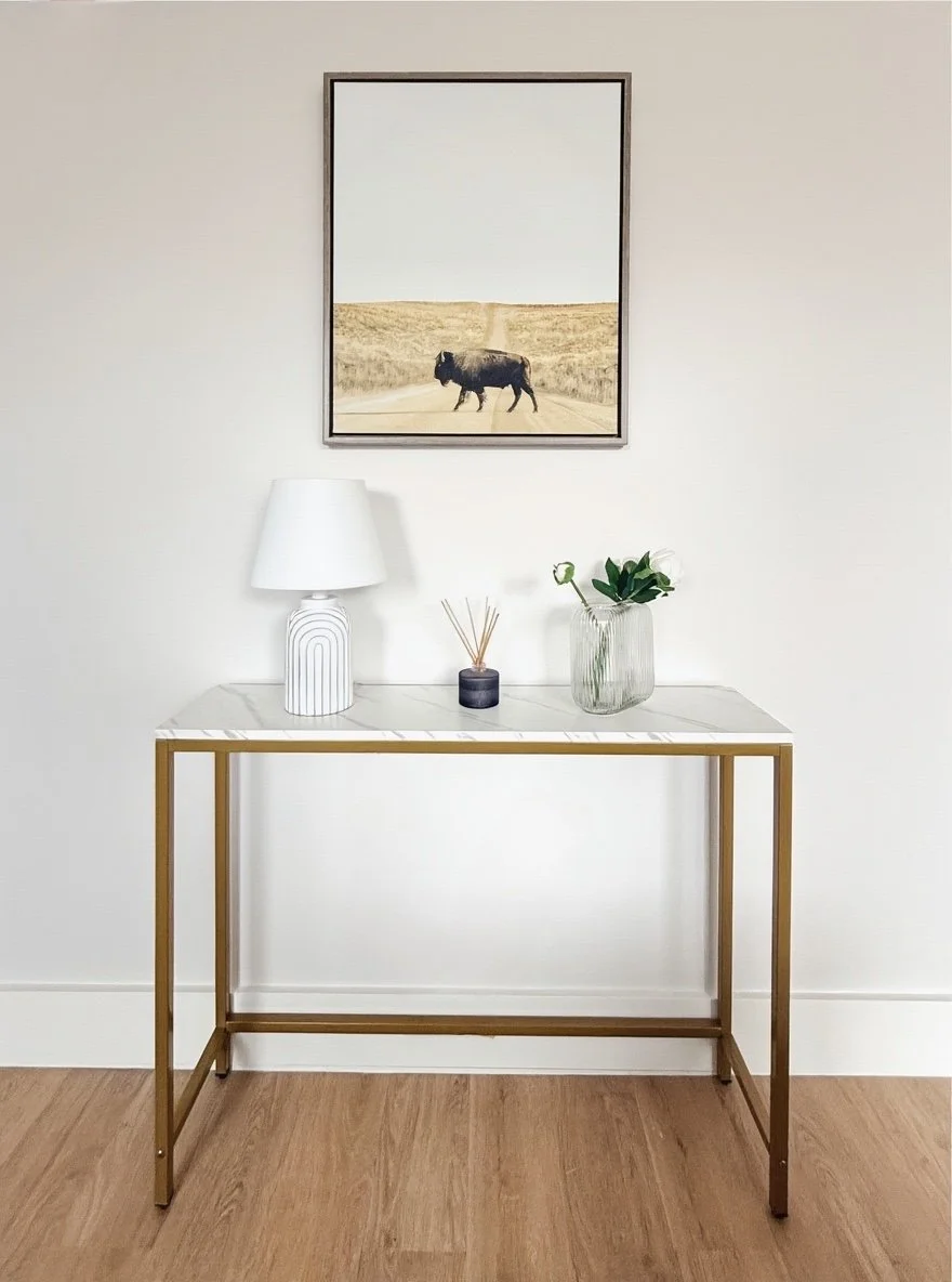 A white console table with a marble top, gold frame, and decorative items including a white lamp, a small black vase with reed diffusers, and a glass vase with green leaves. Above the table hangs a framed picture of a bison on a dirt road in a rural 