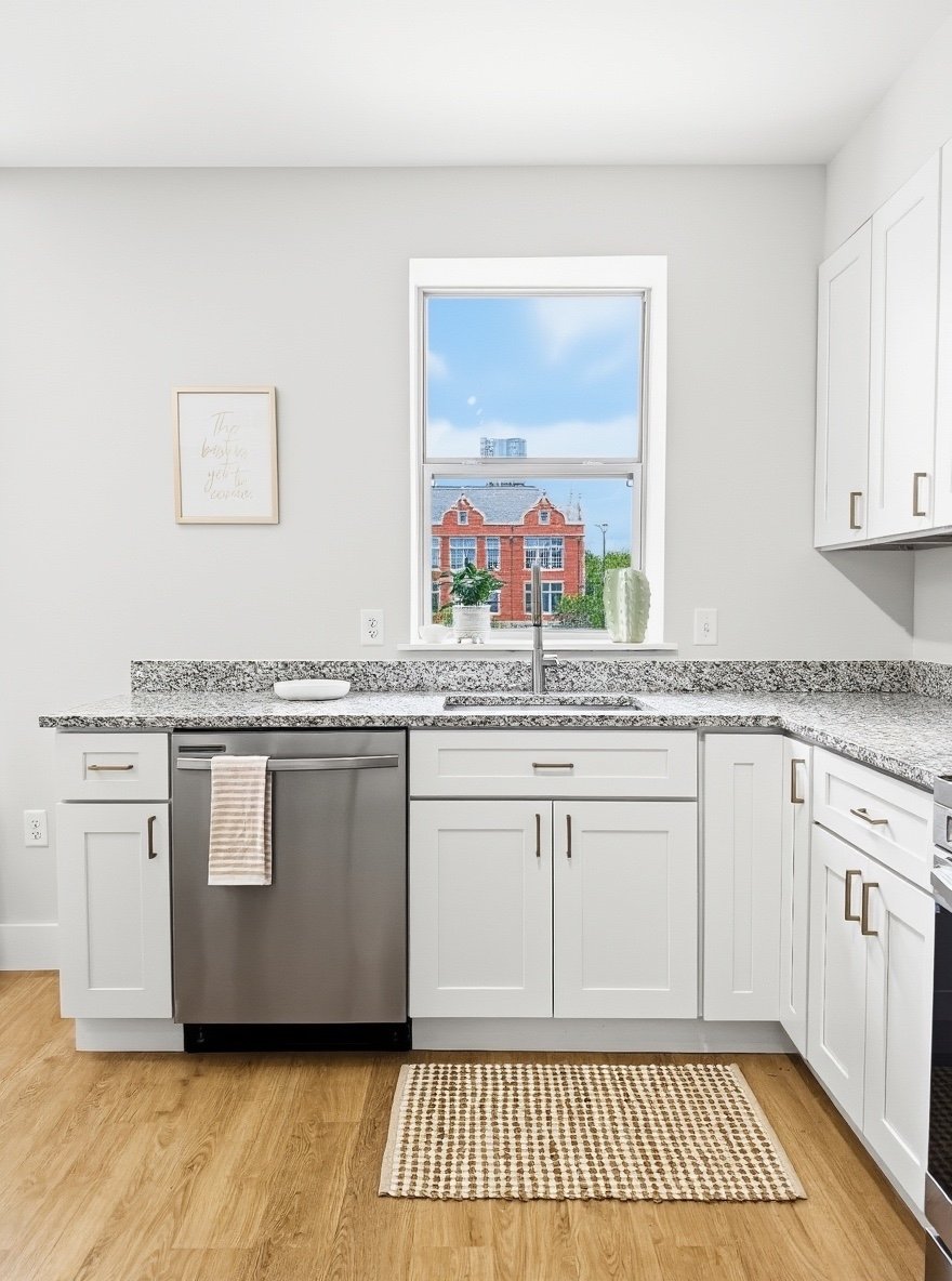 Bright modern kitchen with white cabinets, granite countertops, and a stainless steel dishwasher. A window above the sink offers a view of a brick building and blue sky. Decor includes potted plant and vases on the counter, with a small patterned rug