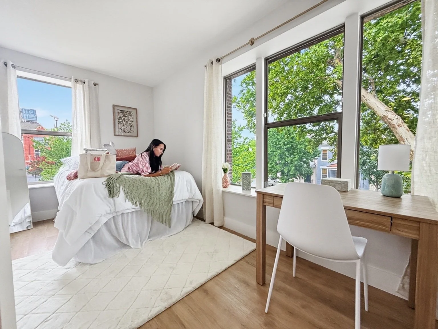 A woman lying on a bed in a bright room with large windows, reading a book. The room has white walls, light wooden flooring, a white rug, a wooden desk with a white chair, a small table lamp, and decorative items on the window sill, with a view of gr