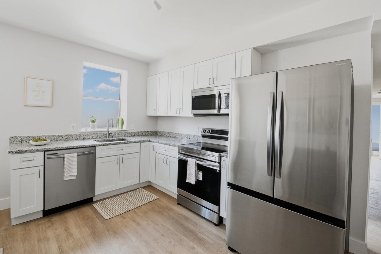 Modern kitchen with white cabinets, stainless steel fridge, stove, microwave, granite countertops, a window with a view of blue sky and clouds, and light wood flooring.