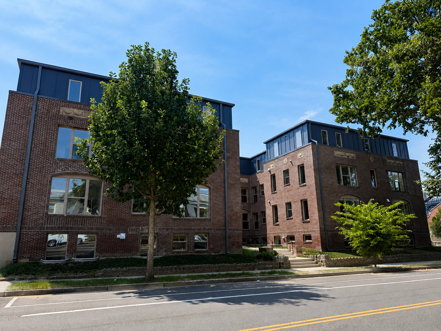 Red brick residential building with black metal roof and windows, two large trees in front, sidewalk, and a street with yellow lines under a clear blue sky.