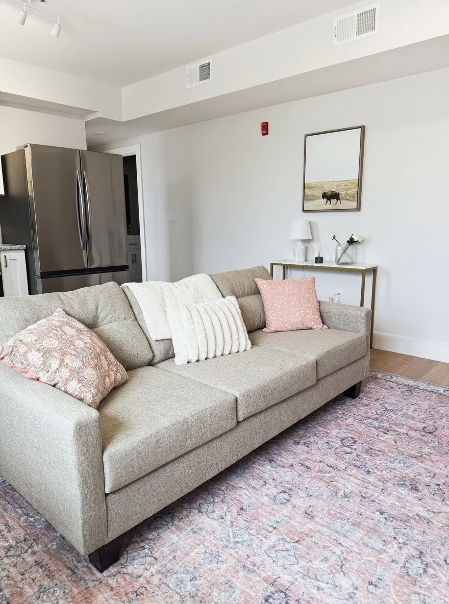 Living room with beige sofa, pink and white pillows, pink patterned rug, white walls, side table with lamp, decorative vase, and framed picture of a cow in a field on the wall, kitchen area with stainless steel refrigerator in background.
