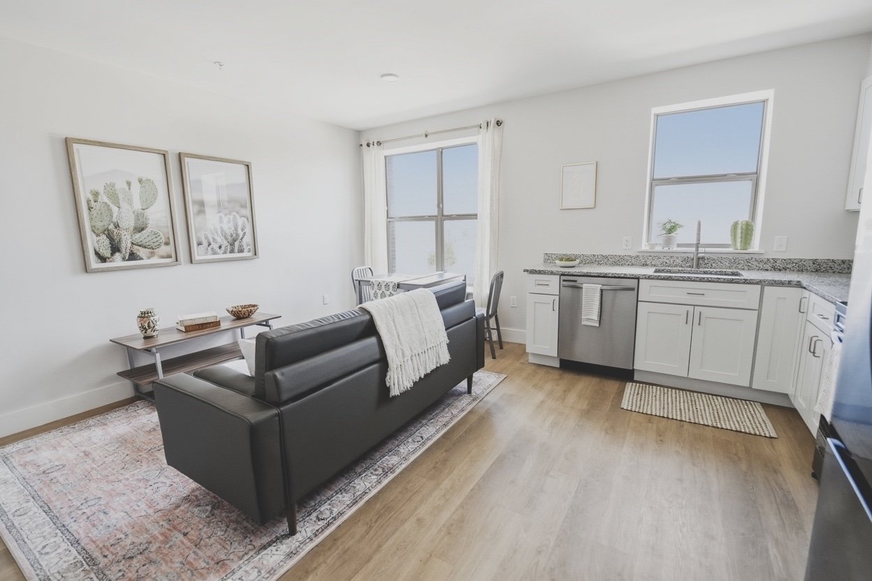 Open-concept living room and kitchen with white walls, wooden flooring, a black leather sofa, a small wooden palm tree stencil art on the wall, a dining table with chairs, large windows with white curtains, and a kitchen with white cabinets and grani