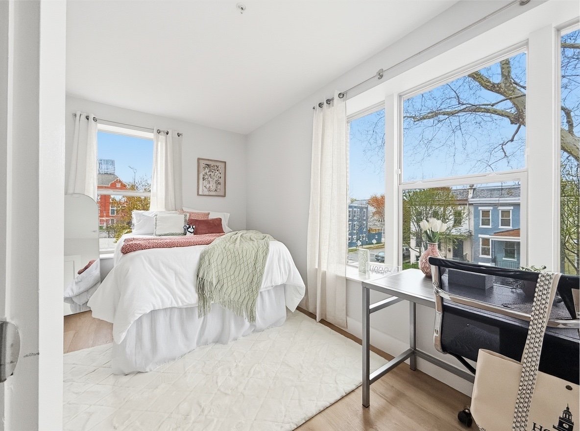 Bright bedroom with white bed, green throw blanket, decorative pillows, large window with views of trees and houses, and a desk with a vase of white flowers.