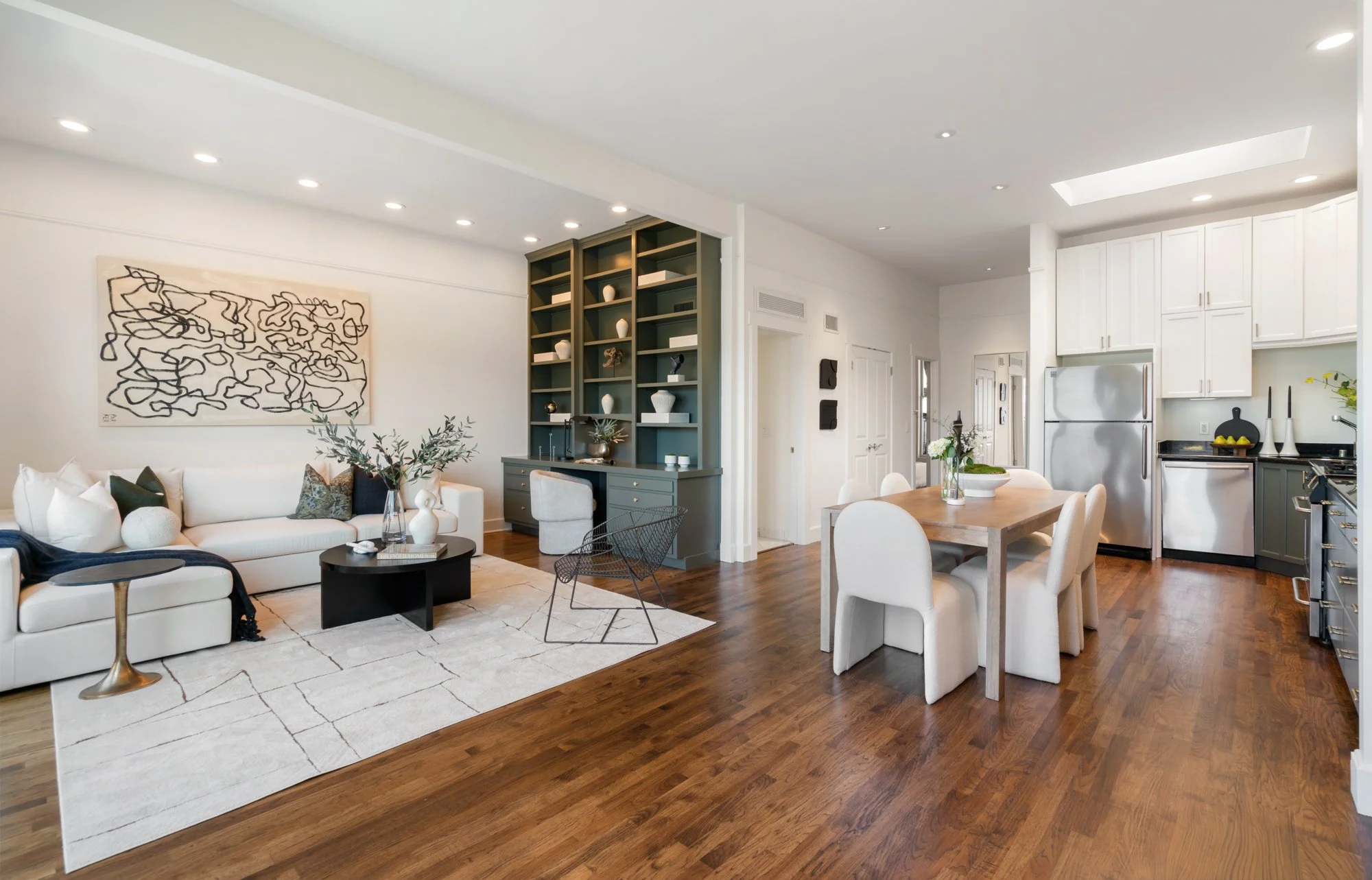 Open-concept living room and kitchen with white walls, hardwood flooring, a gray built-in bookshelf, white kitchen cabinets, and modern furniture including a white sofa, black coffee table, and dining table with white chairs.