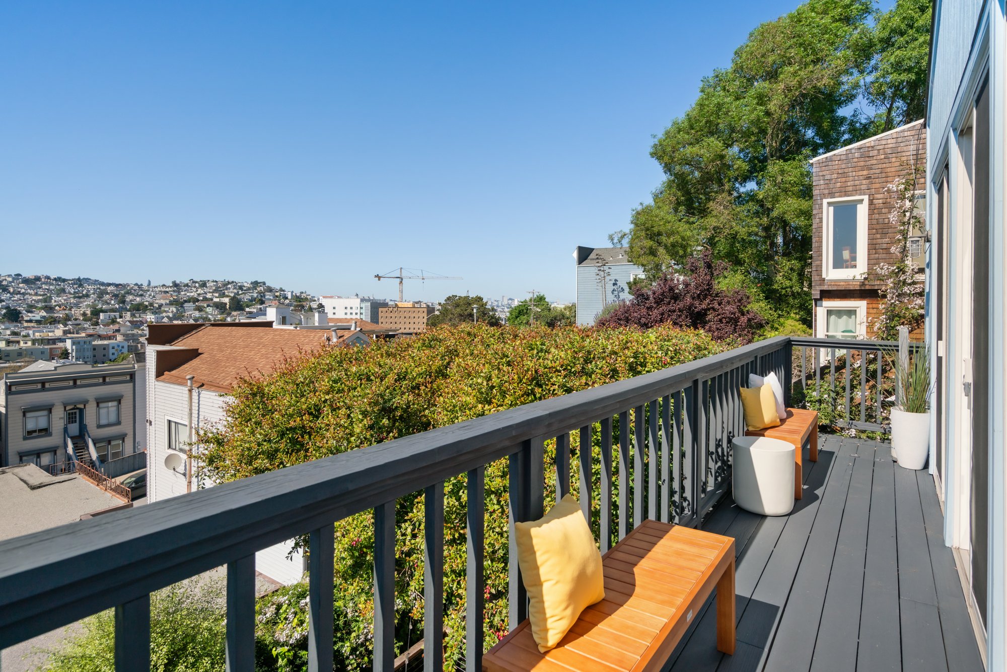 View from a balcony with gray flooring, wooden benches with yellow cushions, a white ottoman, and a large potted plant, overlooking a cityscape with trees, buildings, and a clear blue sky.