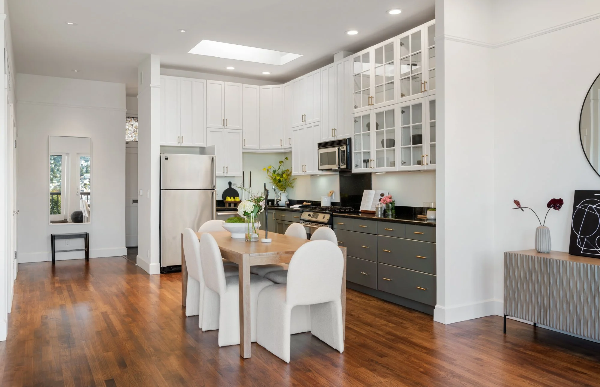 Modern kitchen and dining area with white cabinetry, gray lower cabinets, wooden dining table, six white upholstered chairs, and hardwood floors.
