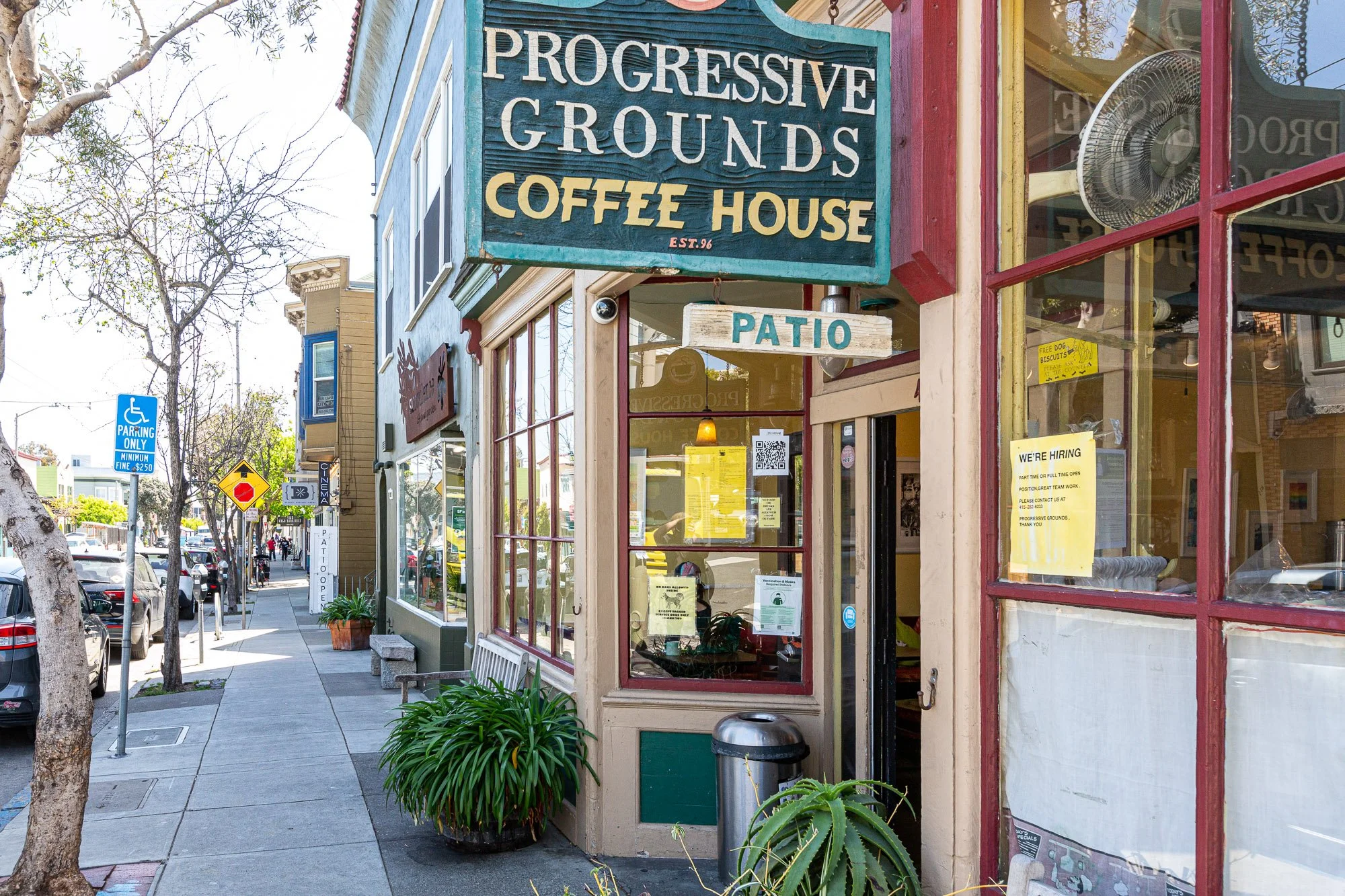 Street view of Progressive Grounds Coffee House with a sign and patio seating. Clear sidewalk, some parked cars, trees, and a blue parking sign in the background.