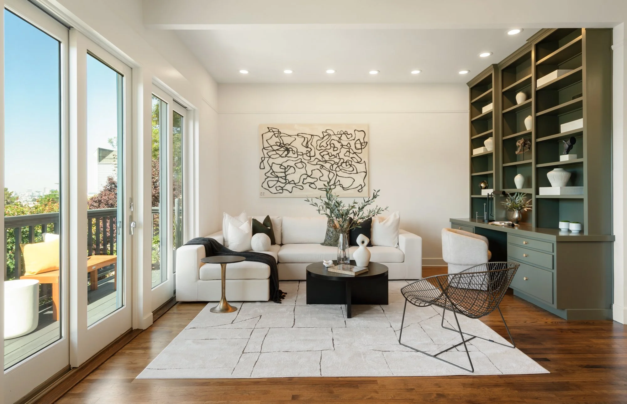 Modern living room with white sofa, green built-in shelves with white vases, black coffee table with decorative objects, black wire chair, sliding glass doors leading to a balcony with outdoor seating, hardwood floors, and recessed ceiling lights.