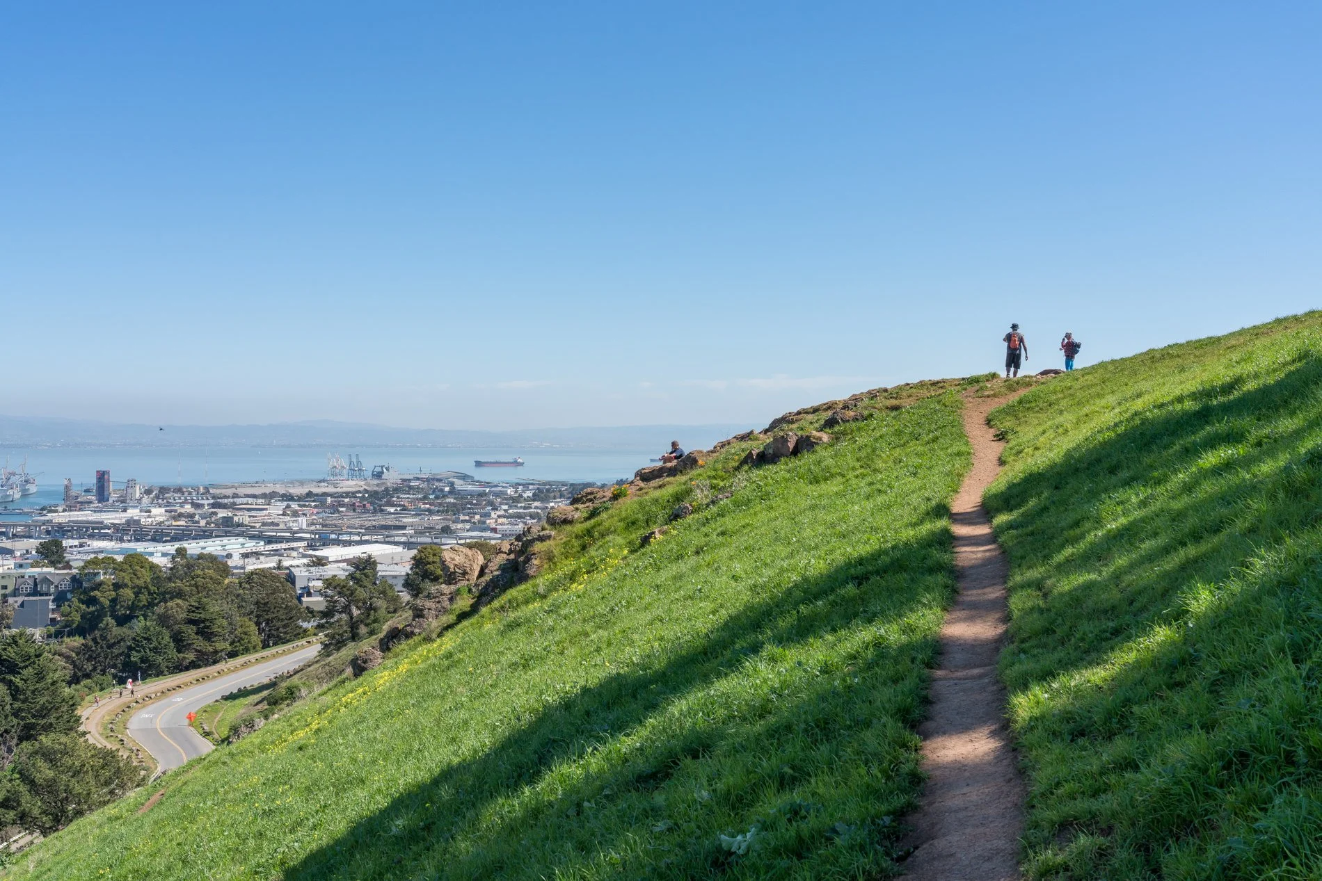 Hiking trail on grassy hillside overlooking a city and harbor with ships, clear blue sky, three hikers in distance, some rocks near the trail.