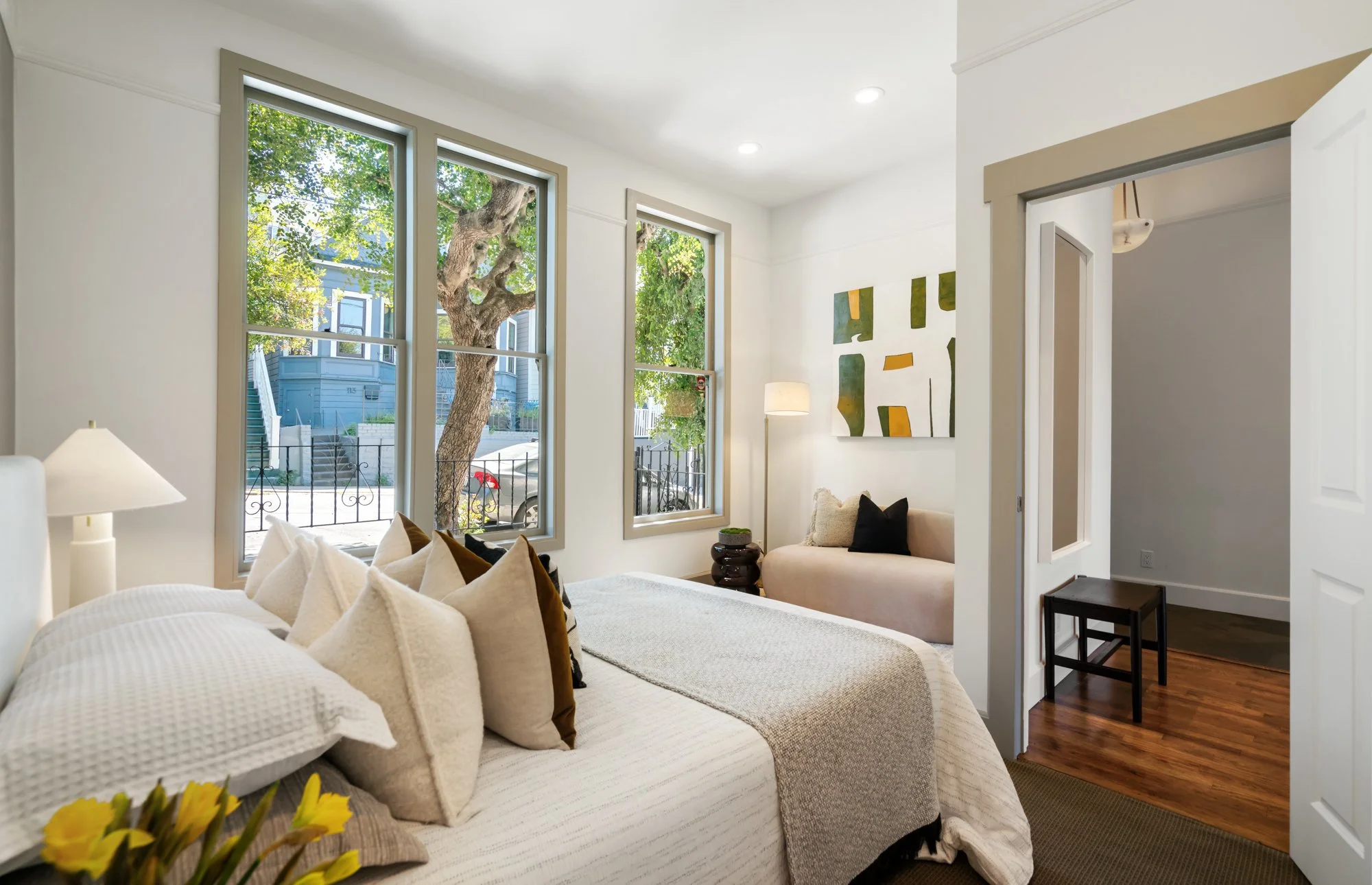 Bright bedroom with two large windows, a bed with many pillows, a small seating area with a beige sofa, a painting on the wall, and a view of a tree outside.