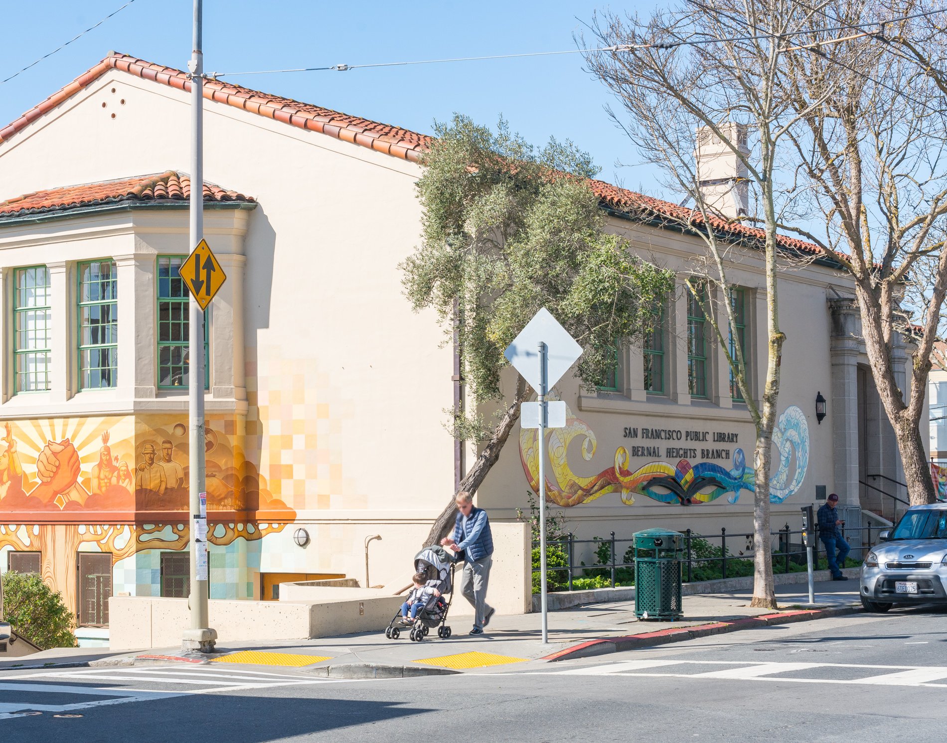 San Francisco Public Library Bernal Heights Branch building with mural art on the exterior wall, sidewalk with a person pushing a stroller, trees, cars, and street signs, clear blue sky.
