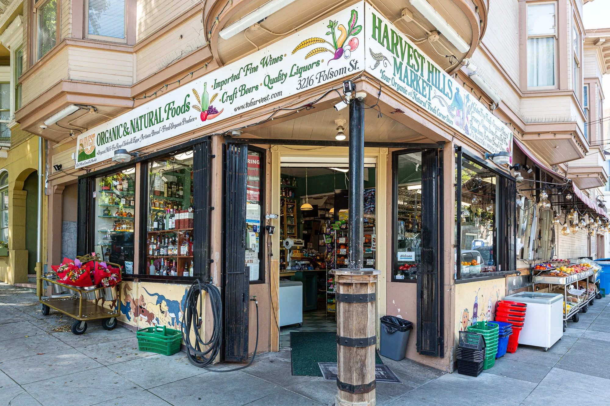 Corner view of a small natural foods and market shop on a city street, with produce and flower displays outside and signage advertising organic foods, wines, craft beers, and liquors.