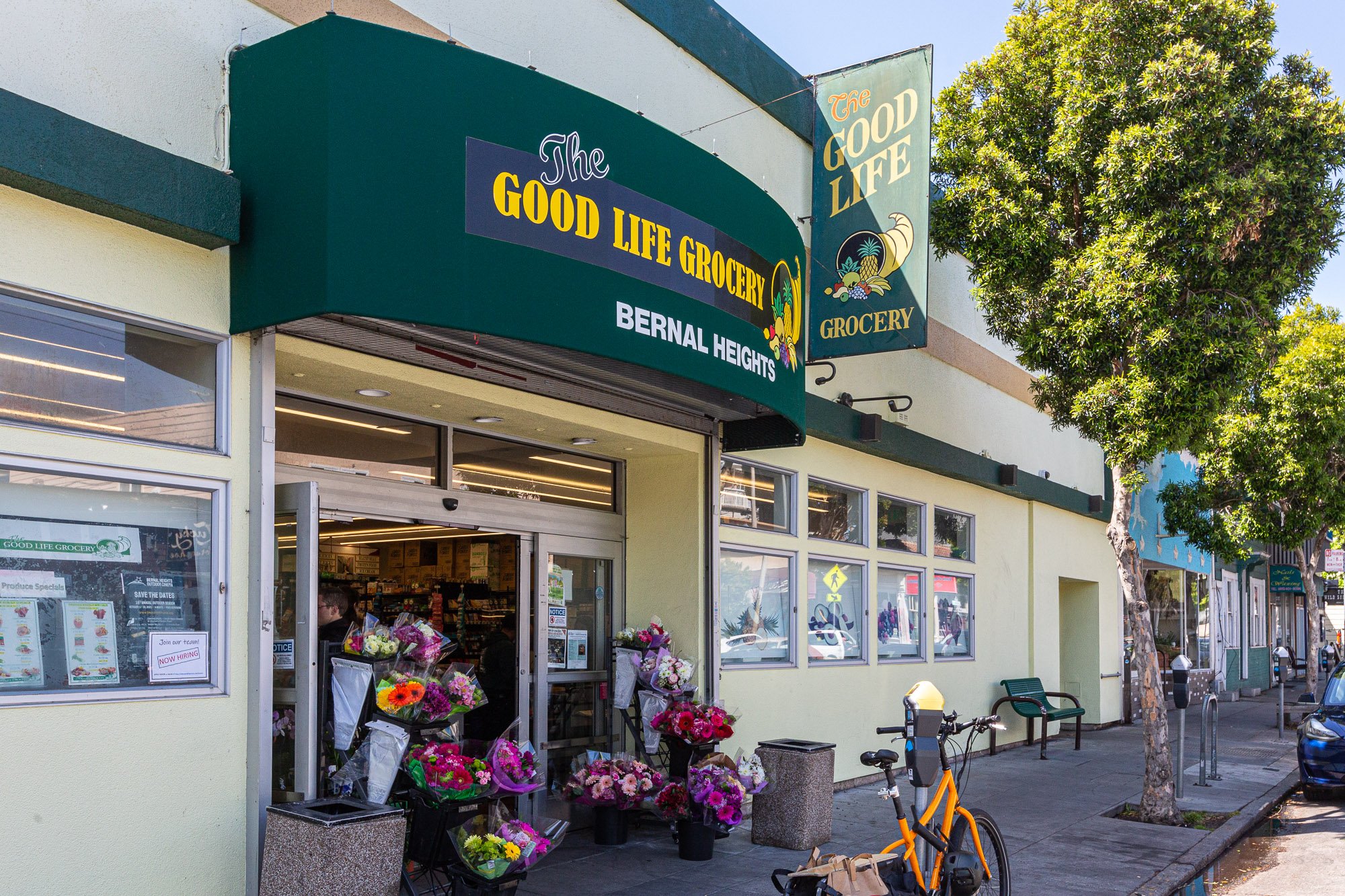  storefront of The Good Life Grocery with flower bouquets outside and a bicycle parked in front, trees lining the sidewalk on a sunny day.