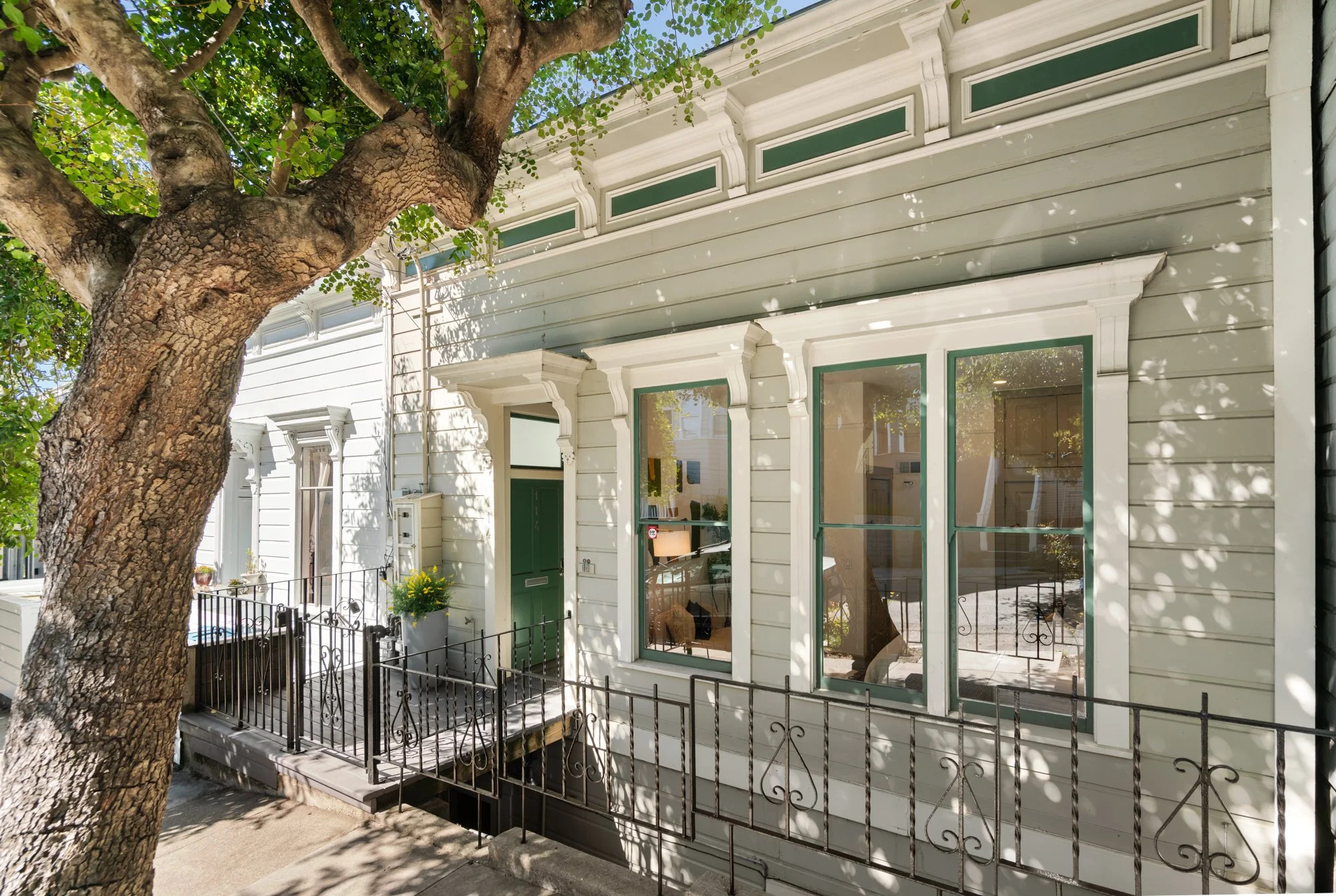 Front view of a vintage white house with green window frames, a small porch, and a large tree in front casting shadows on the house.