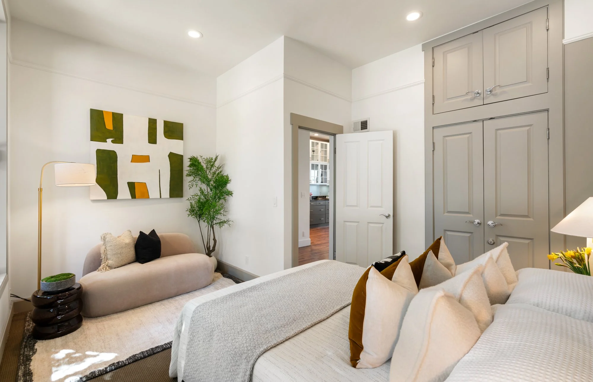 Interior view of a bedroom with a bed, pillows, a beige loveseat with pillows, a floor lamp, a potted plant, and artwork on the wall. An open door reveals a hallway and part of a kitchen.