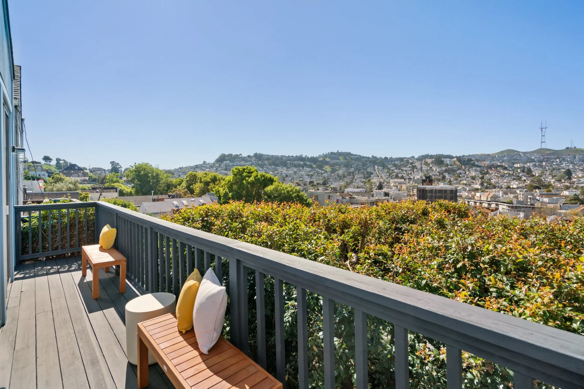 A wooden balcony with seating pillows overlooking a lush cityscape with rolling hills and communication towers under a clear blue sky.