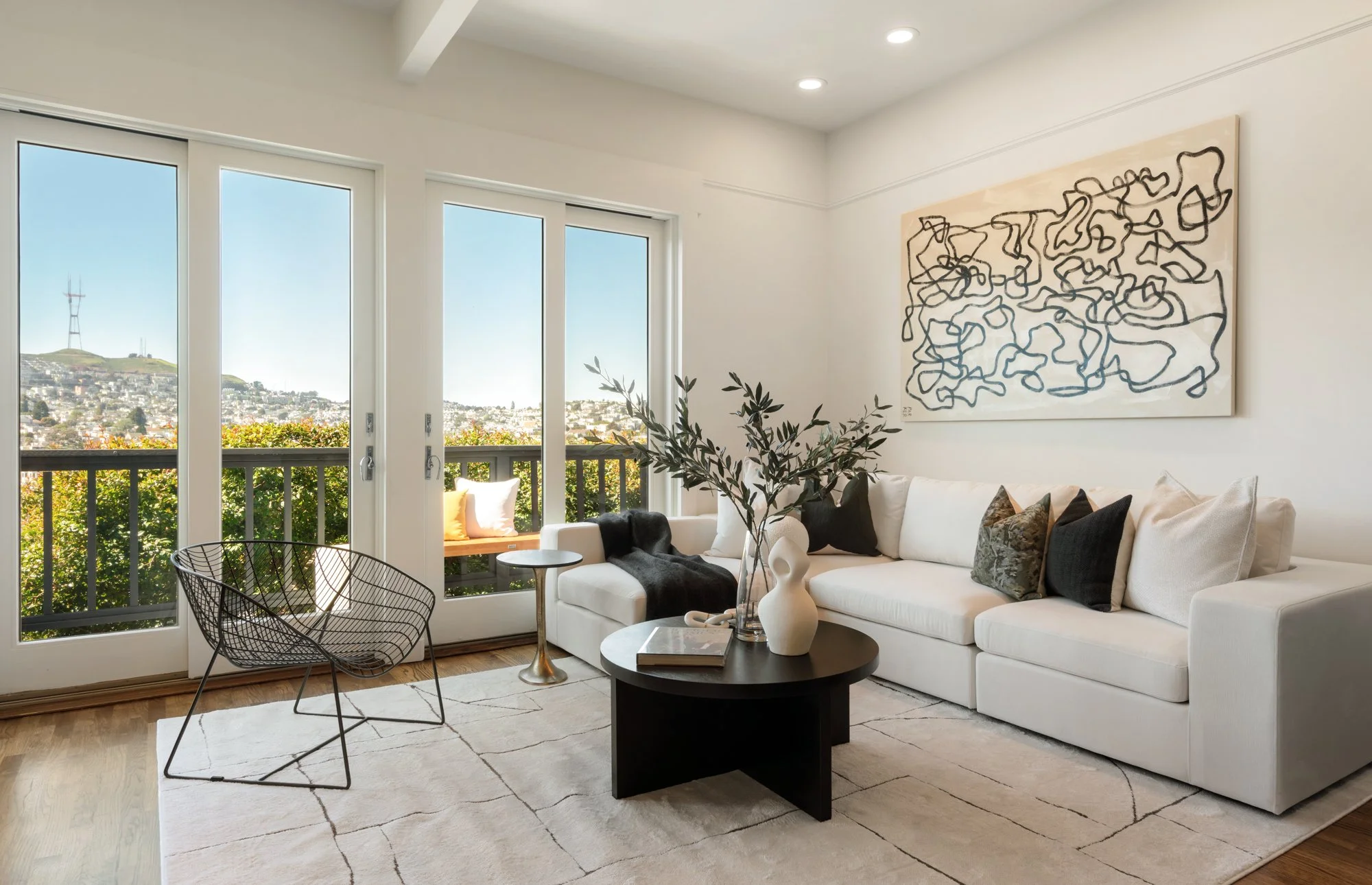 Bright living room with white walls and a large white sectional sofa adorned with several pillows. A black abstract line art painting hangs above the sofa. A black round coffee table with white and black vases and a book sits on a beige area rug. To 