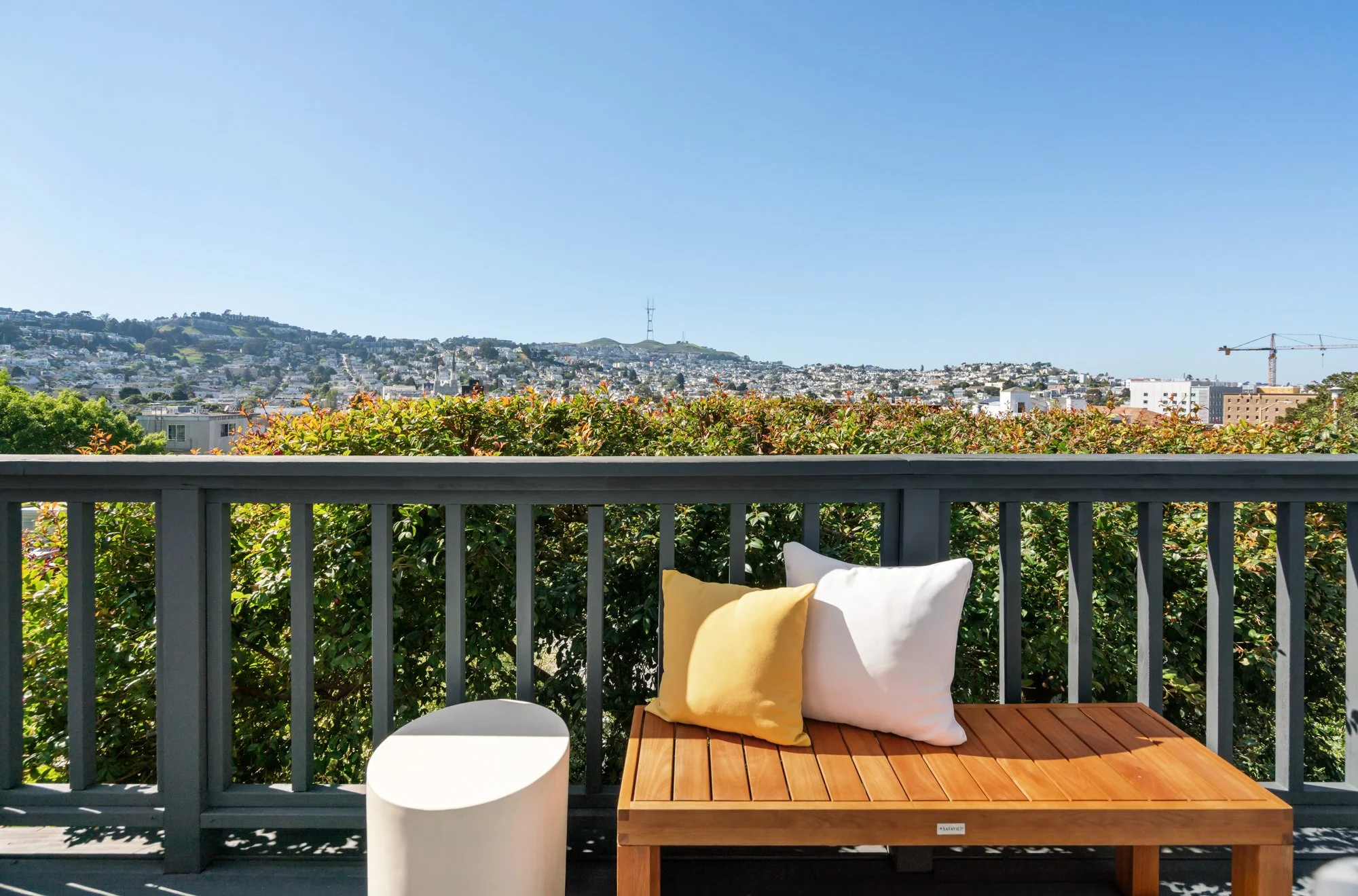 View of a city skyline from a balcony with a wooden bench, two pillows (yellow and white), and a small white table. Green bushes and a clear blue sky are visible.